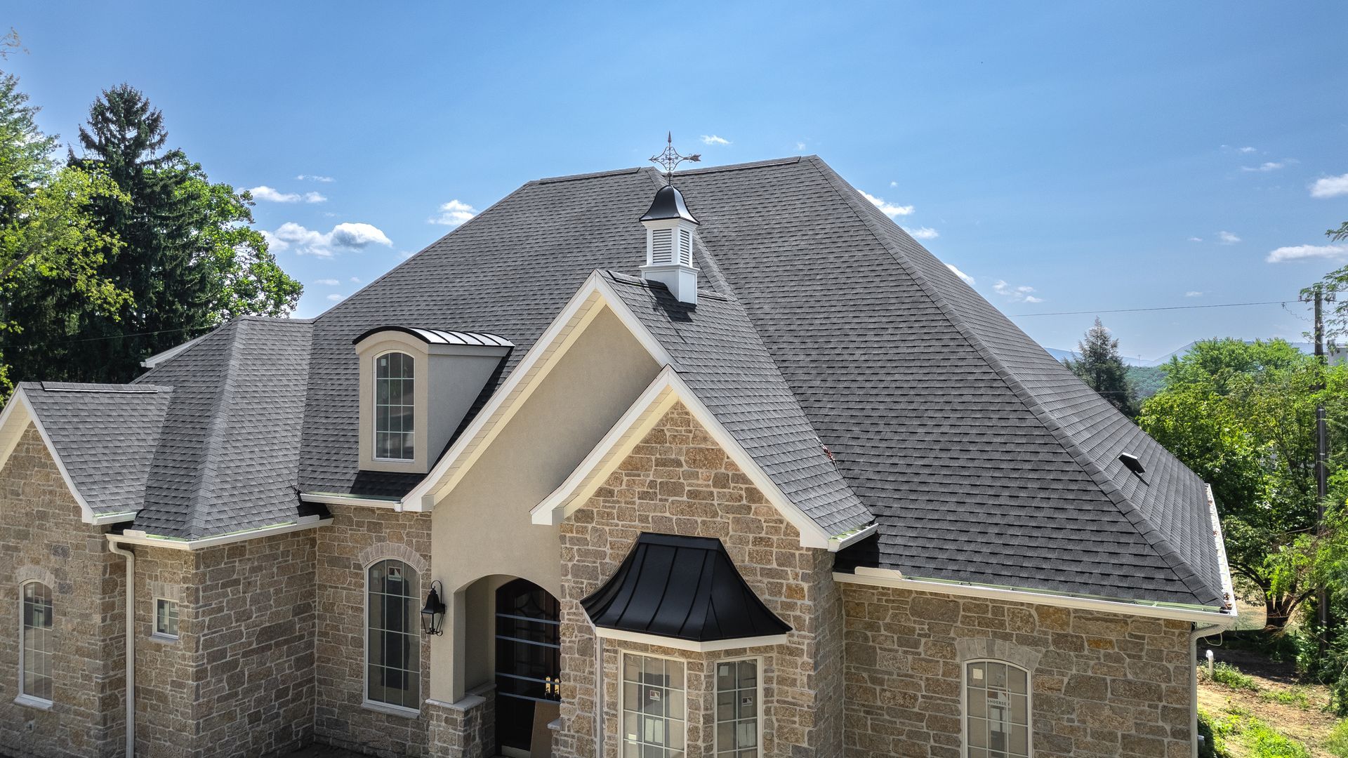 Stone house with dark shingled roof, dormers, and a weathervane against a blue sky.