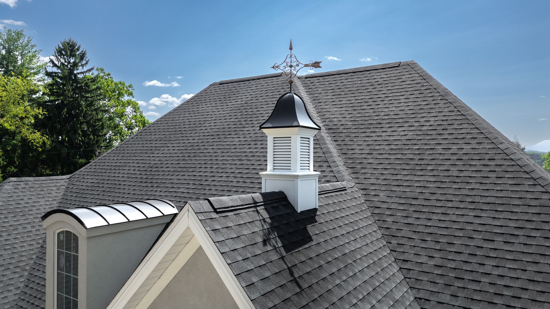 View of a house roof with a weathervane, dormer window, and asphalt shingles. Blue sky and trees in background.