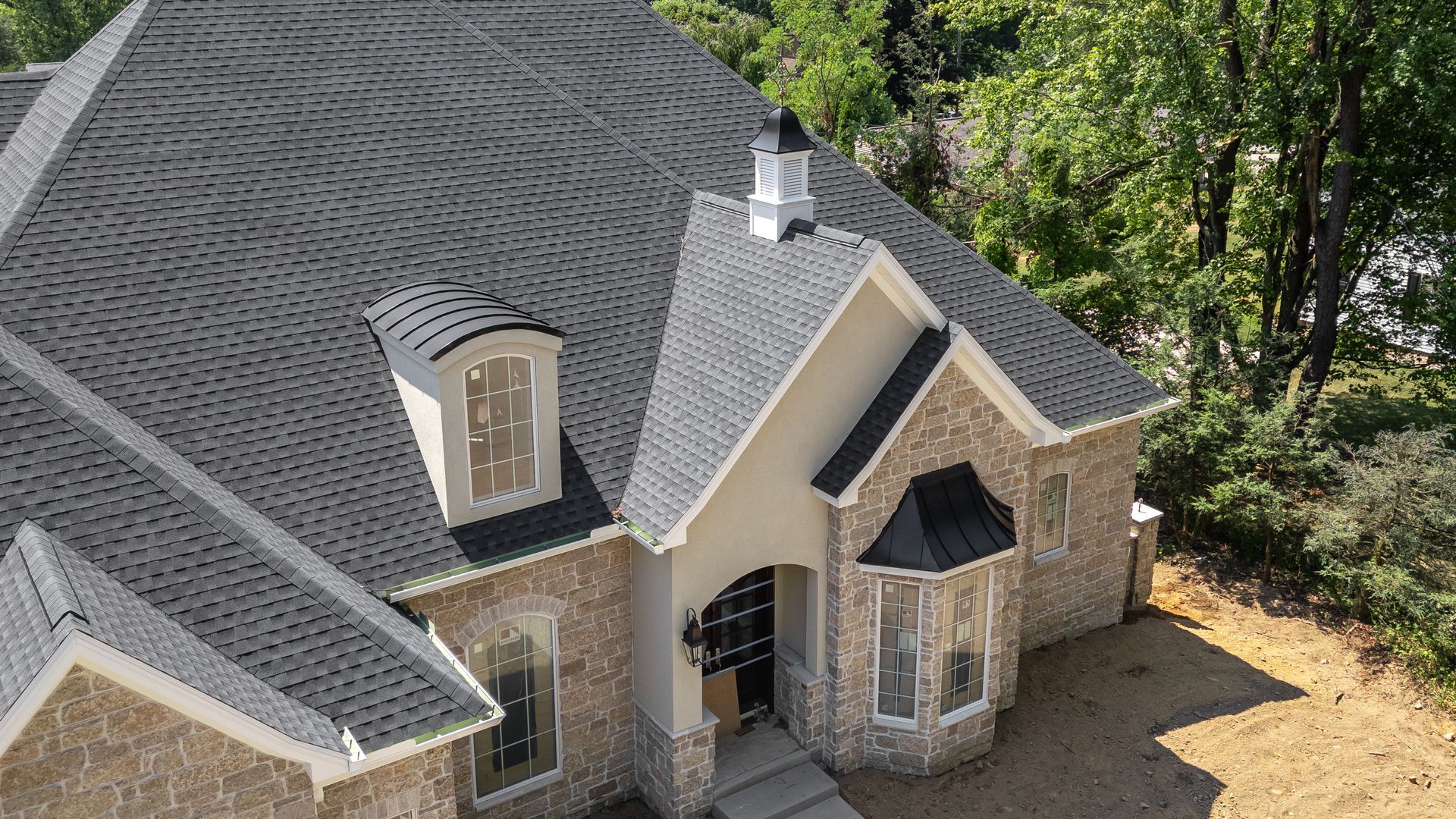 Stone and brick house with gray shingle roof, dormers, and entry, set among trees.