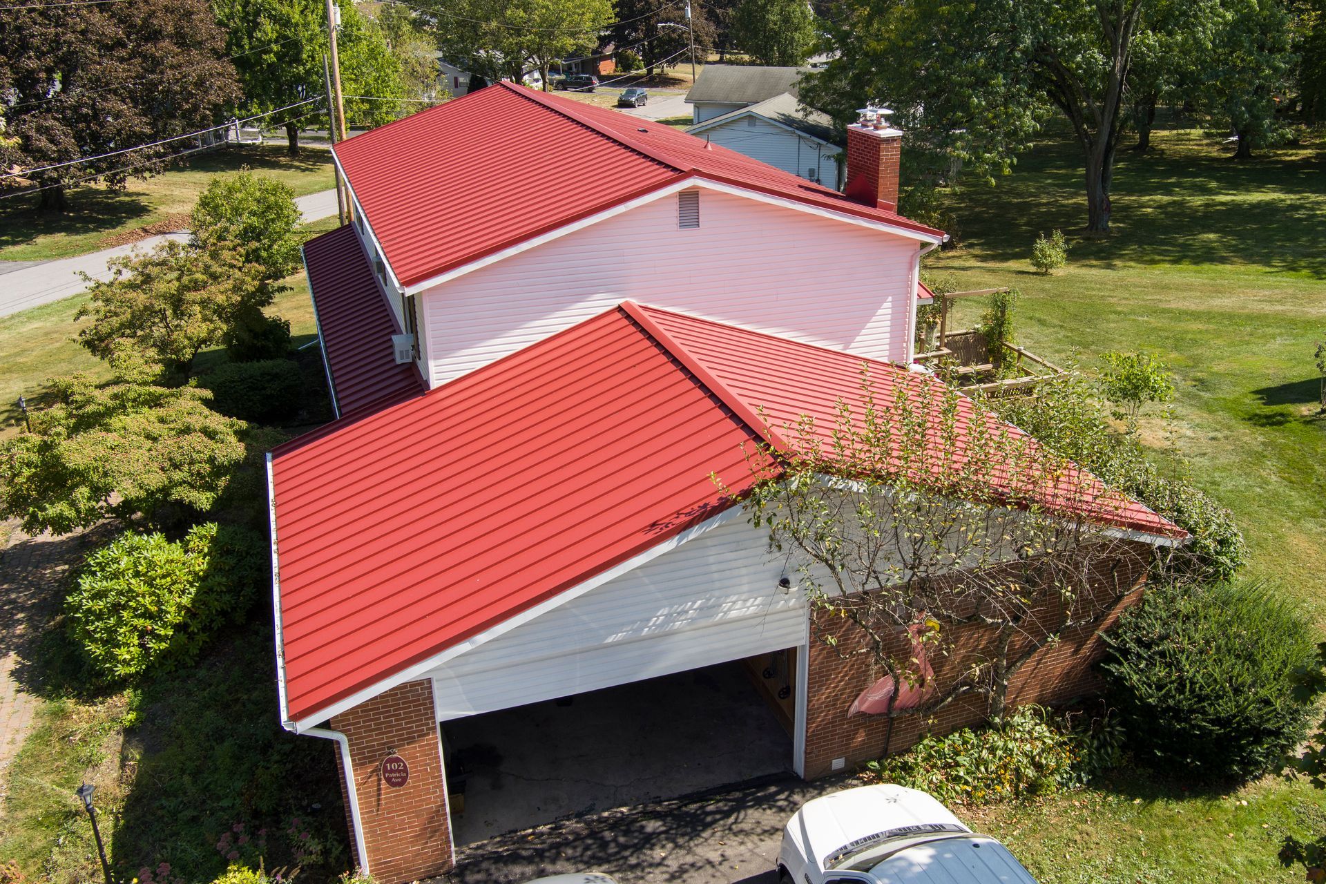 An aerial view of a two-story house with a distinct, bright red metal roof, surrounded by trees and a green lawn.
