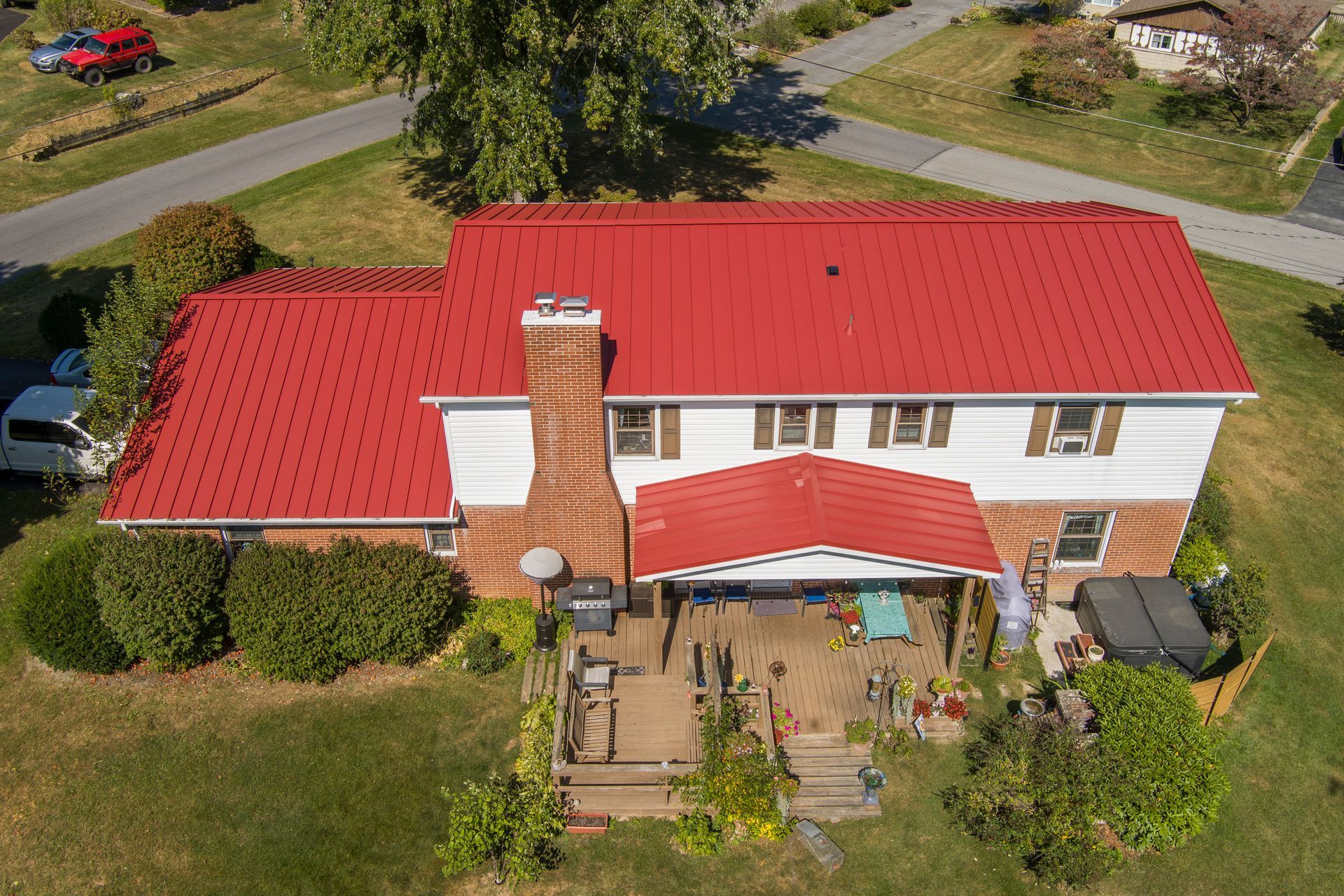 Aerial view of a two-story house with a vibrant red metal roof, a brick chimney, a back deck, and surrounding yard.