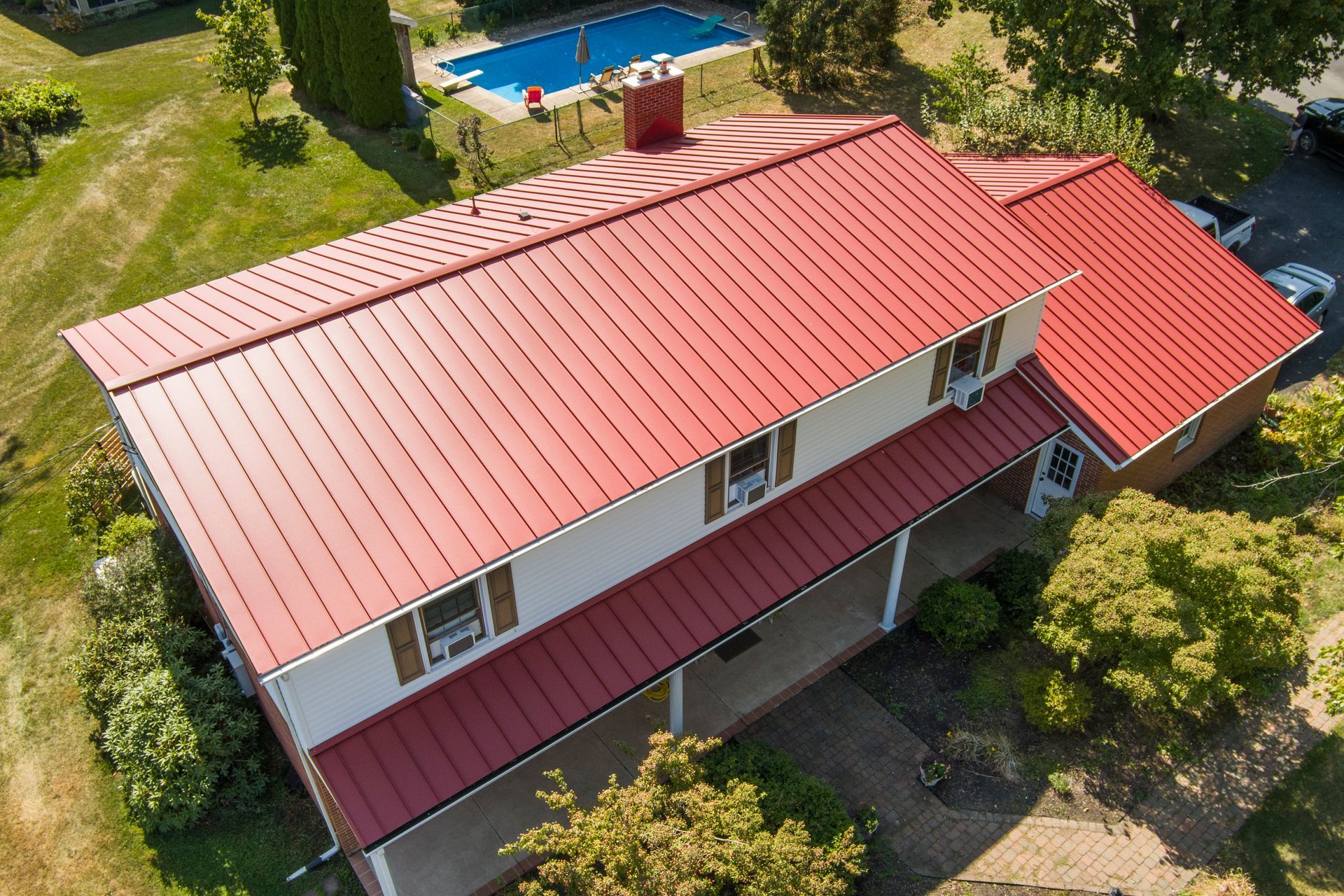 A high-angle view of a two-story house with a prominent red metal roof, white siding, and a backyard swimming pool.