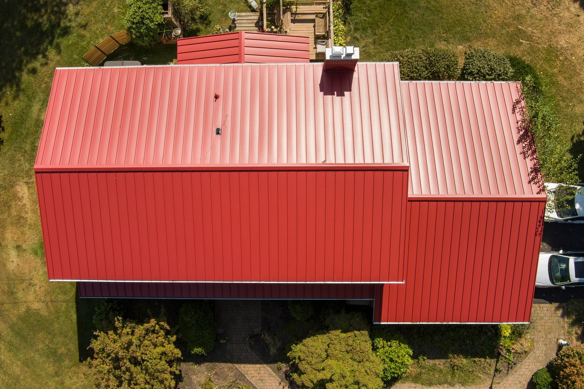 An aerial view of a house with a vibrant red metal roof surrounded by green trees and lawn.