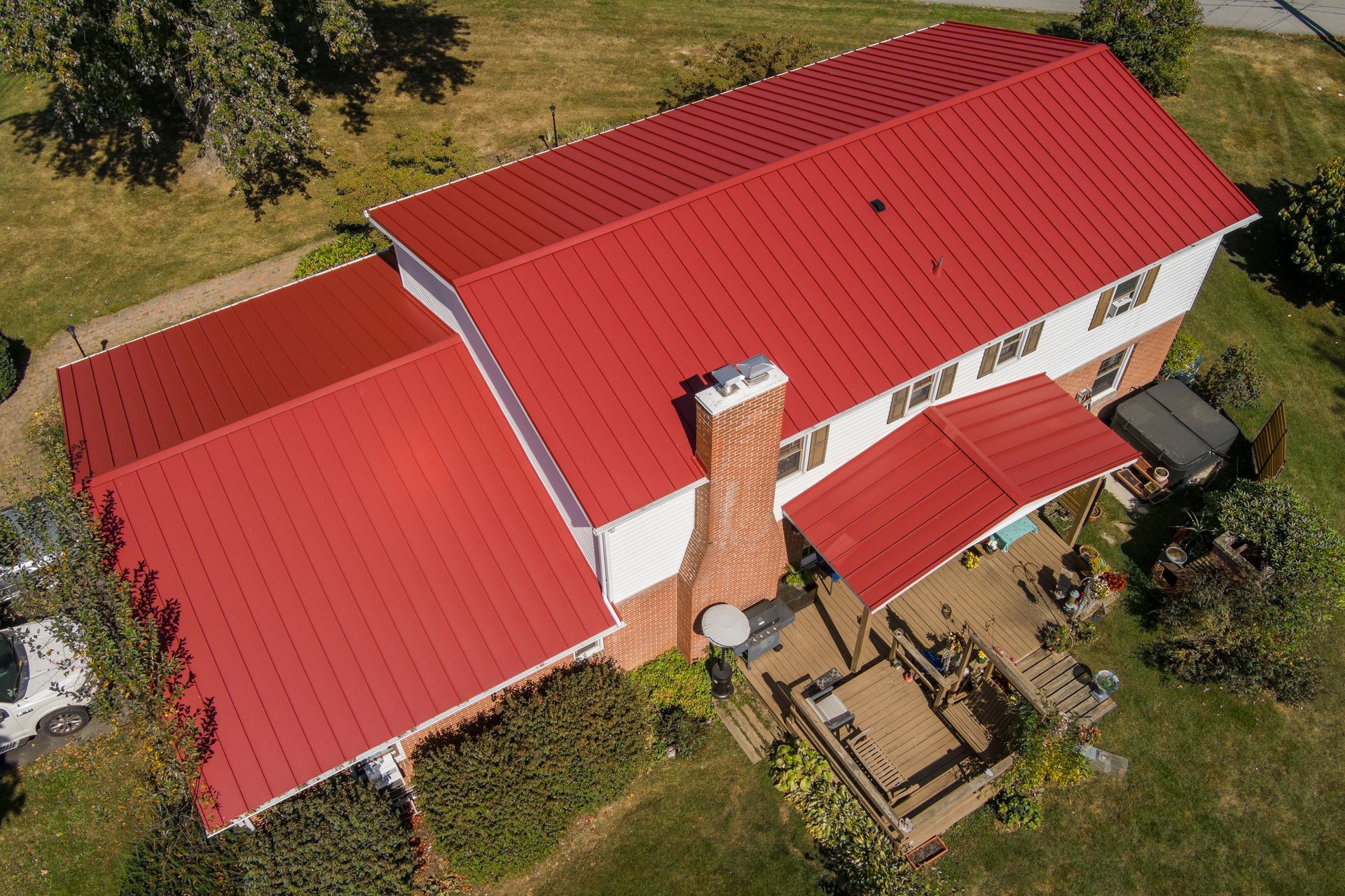 An aerial view of a two-story home with a vibrant red metal roof, a brick chimney, and a wooden deck in a grassy yard.