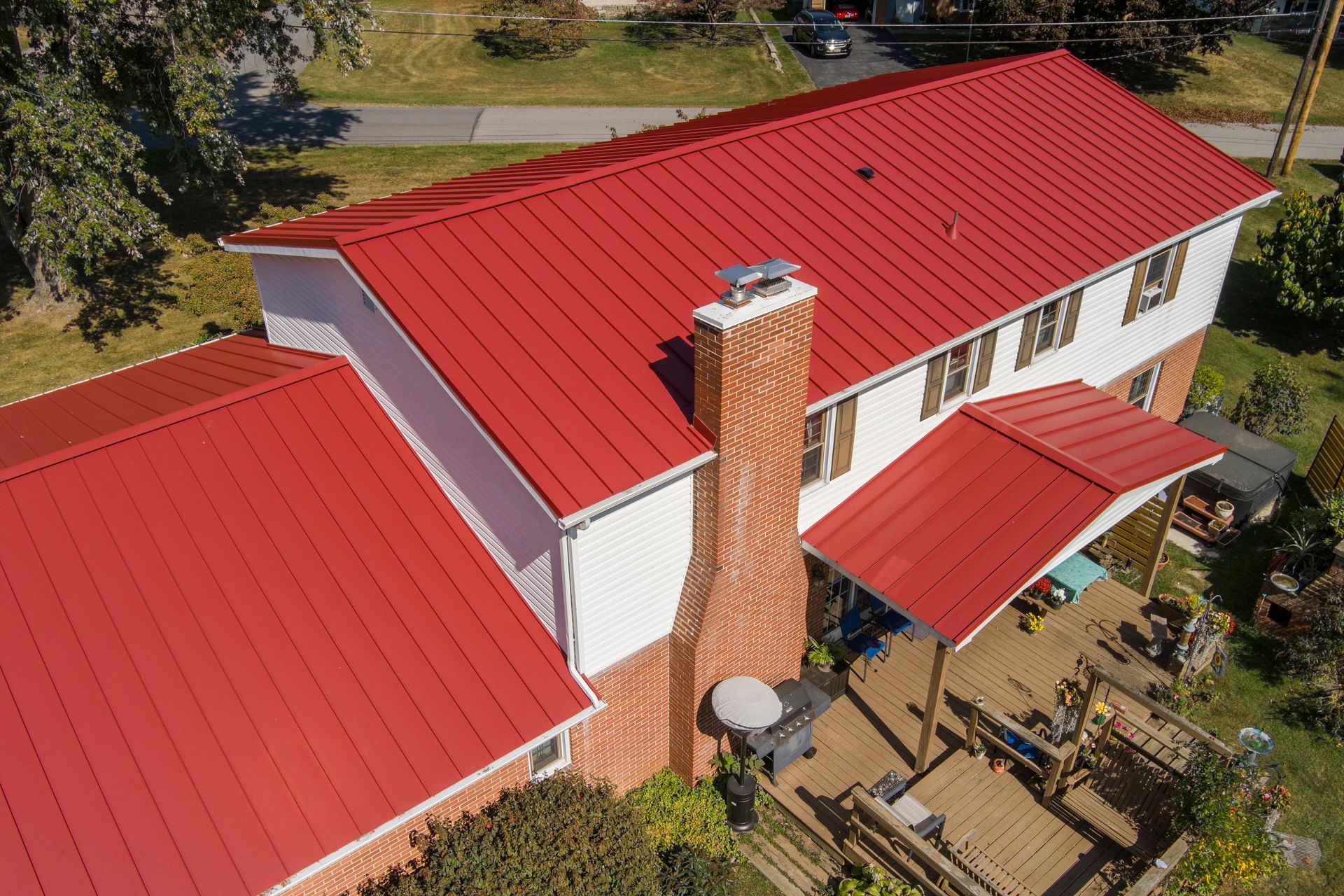Aerial view of a white house with a vibrant red metal roof, a brick chimney, and a wooden deck in a sunny yard.