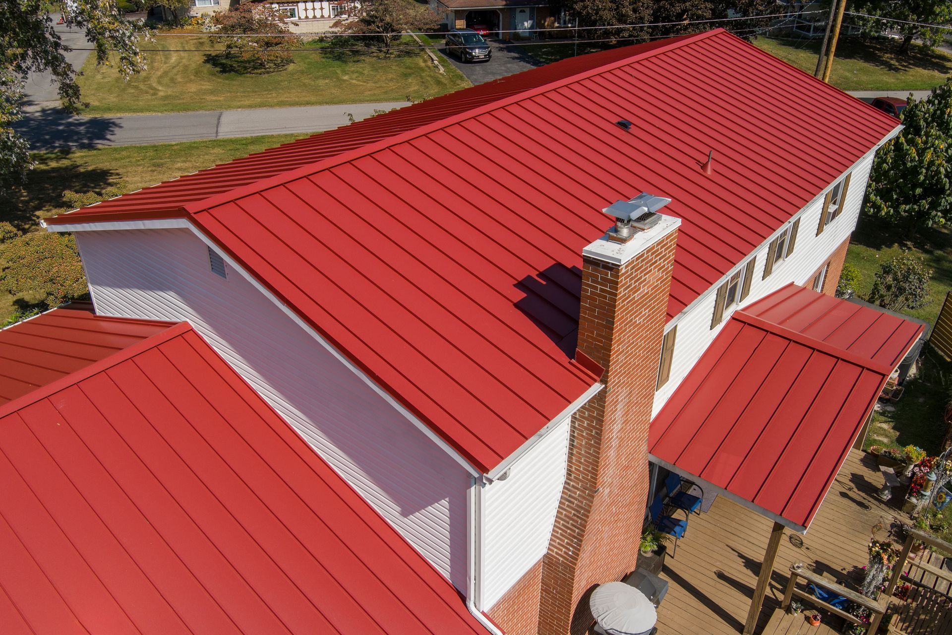 An aerial view of a white residential house featuring a vibrant red metal roof and a brick chimney.