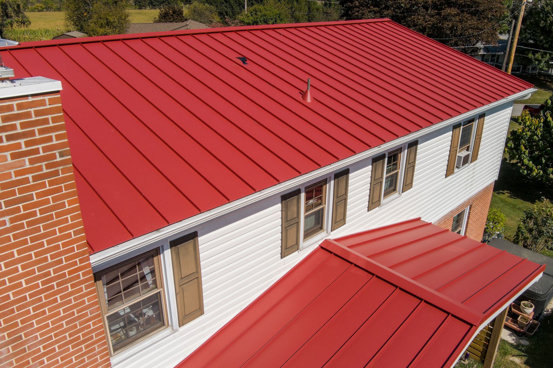 A high-angle view of a white two-story house with a vibrant red standing seam metal roof and a brick chimney.
