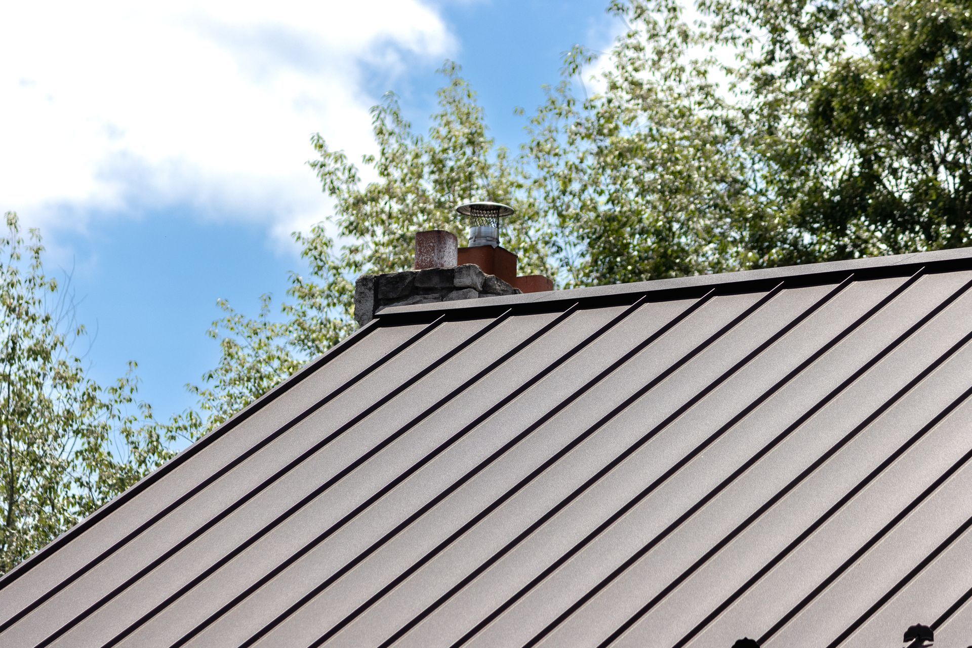Brown metal roof with chimney against a blue sky and trees.