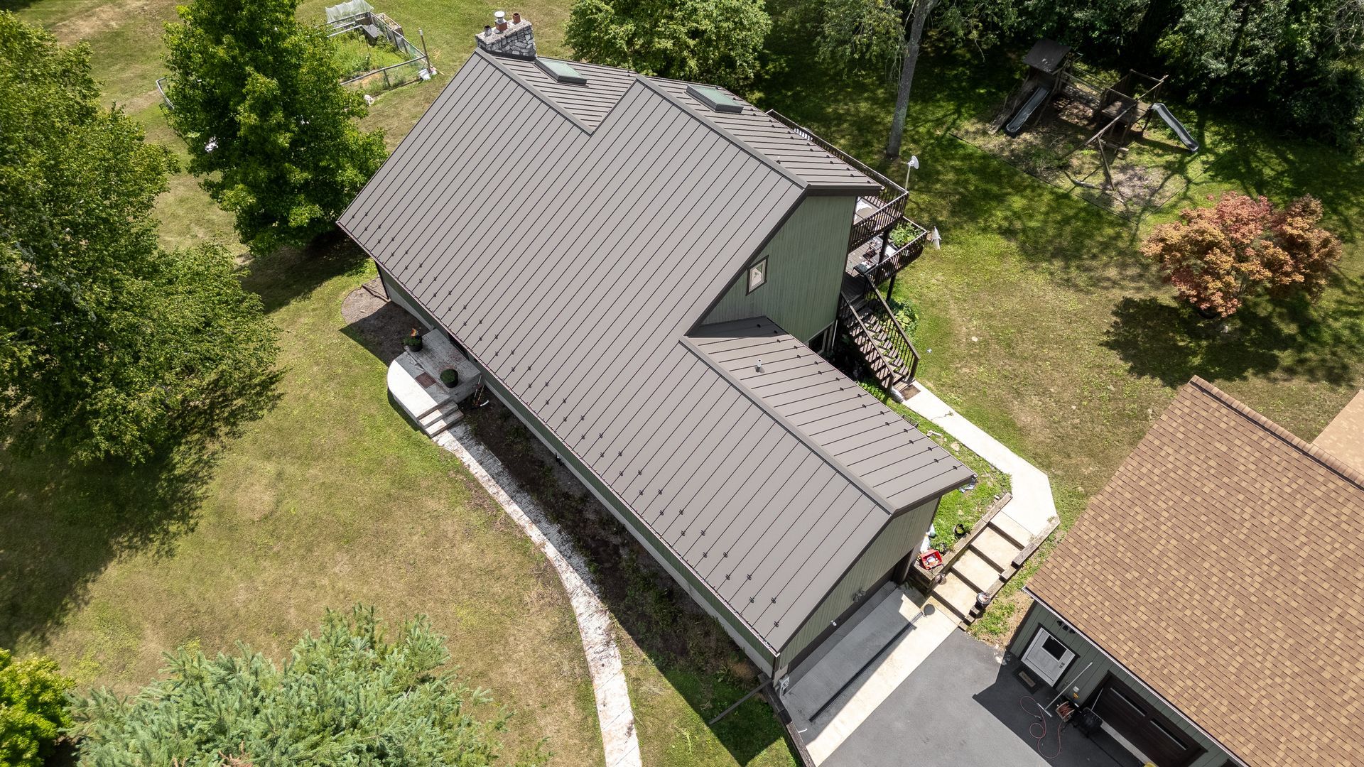 Aerial view of a house with a gray roof, surrounded by green grass and trees; driveway to the right.