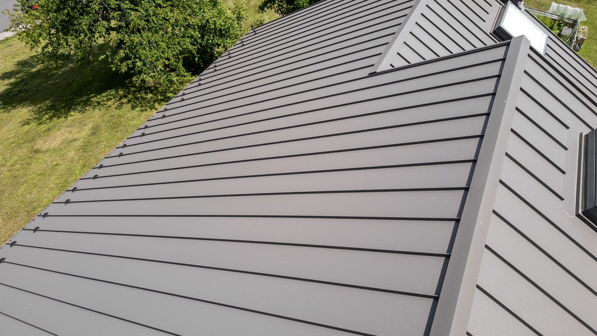 Gray metal roof on a house, with parallel lines and a corner visible. Green grass and tree are on the side.