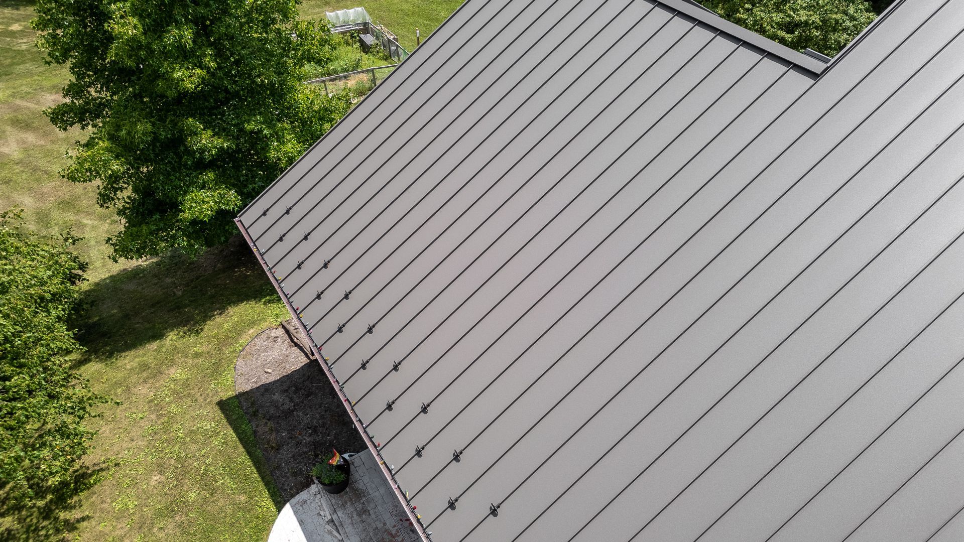 Gray metal roof of a building with a window, viewed from above. Green trees and grass in the background.