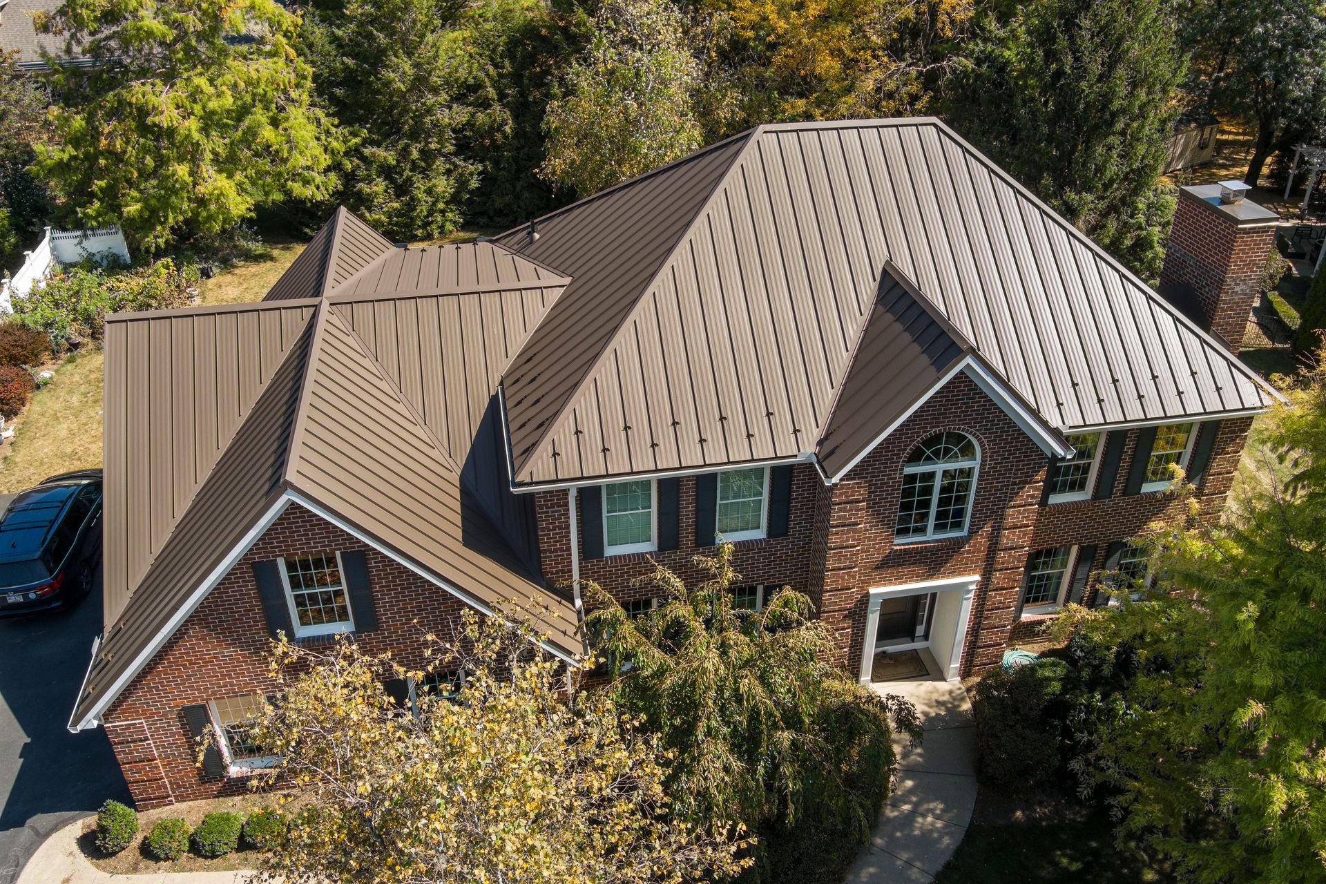 An aerial view of a two-story brick house with a dark brown, textured metal roof, surrounded by trees on a sunny day.