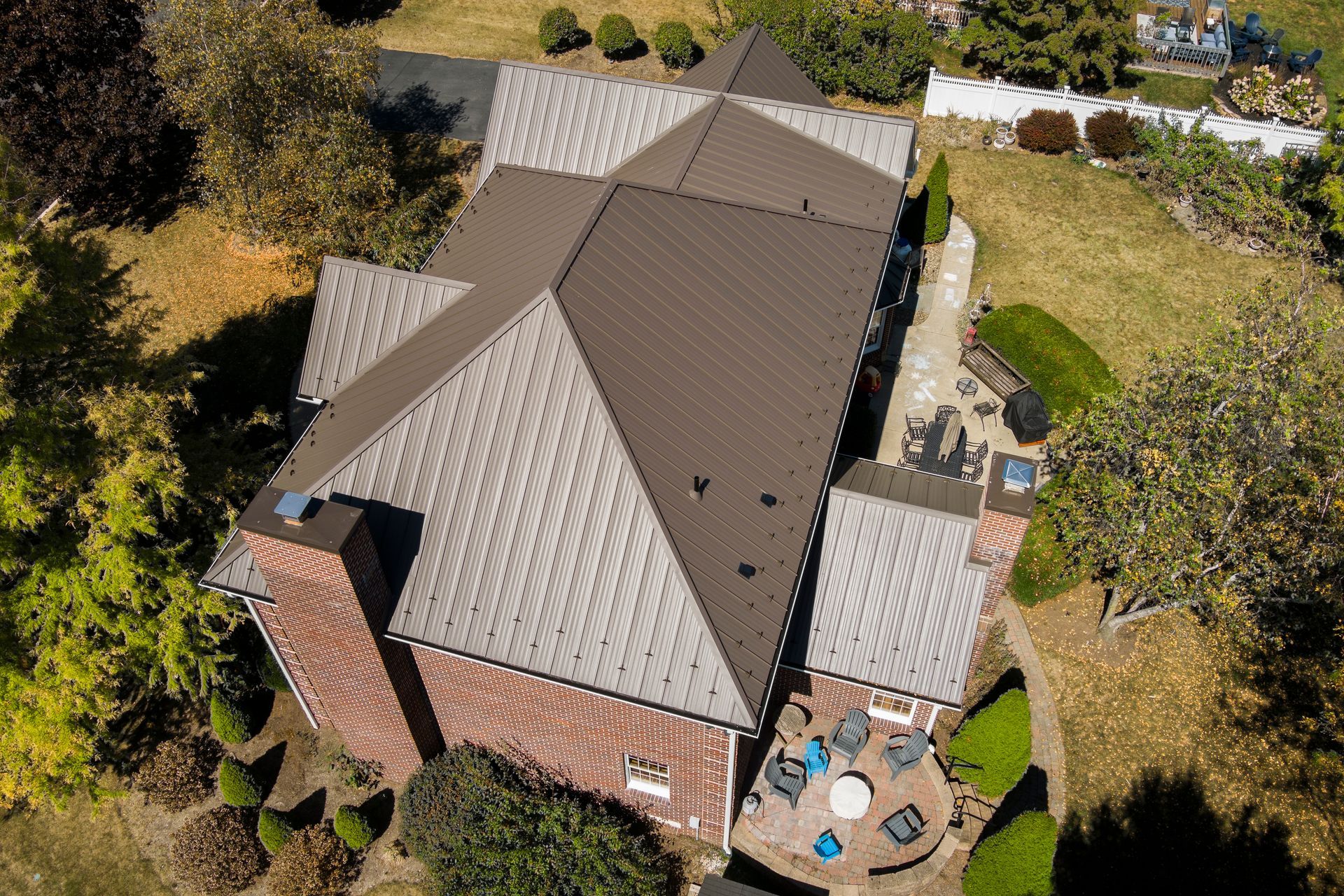 Aerial view of a house with a brown metal roof, brick chimney, and a stone patio area with chairs in a backyard.