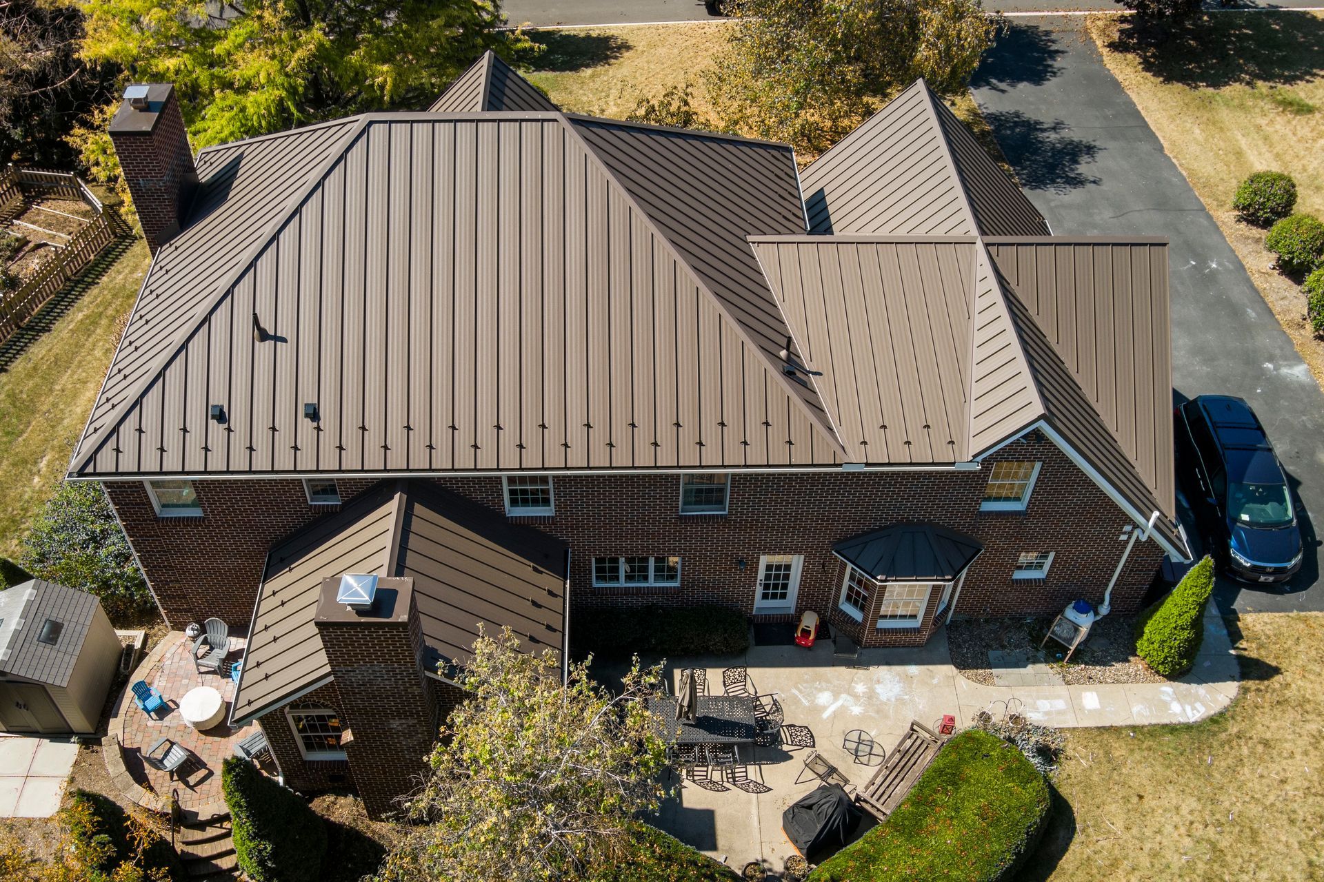 An aerial view of a two-story brick home with a brown metal roof, surrounded by a patio and a paved driveway.