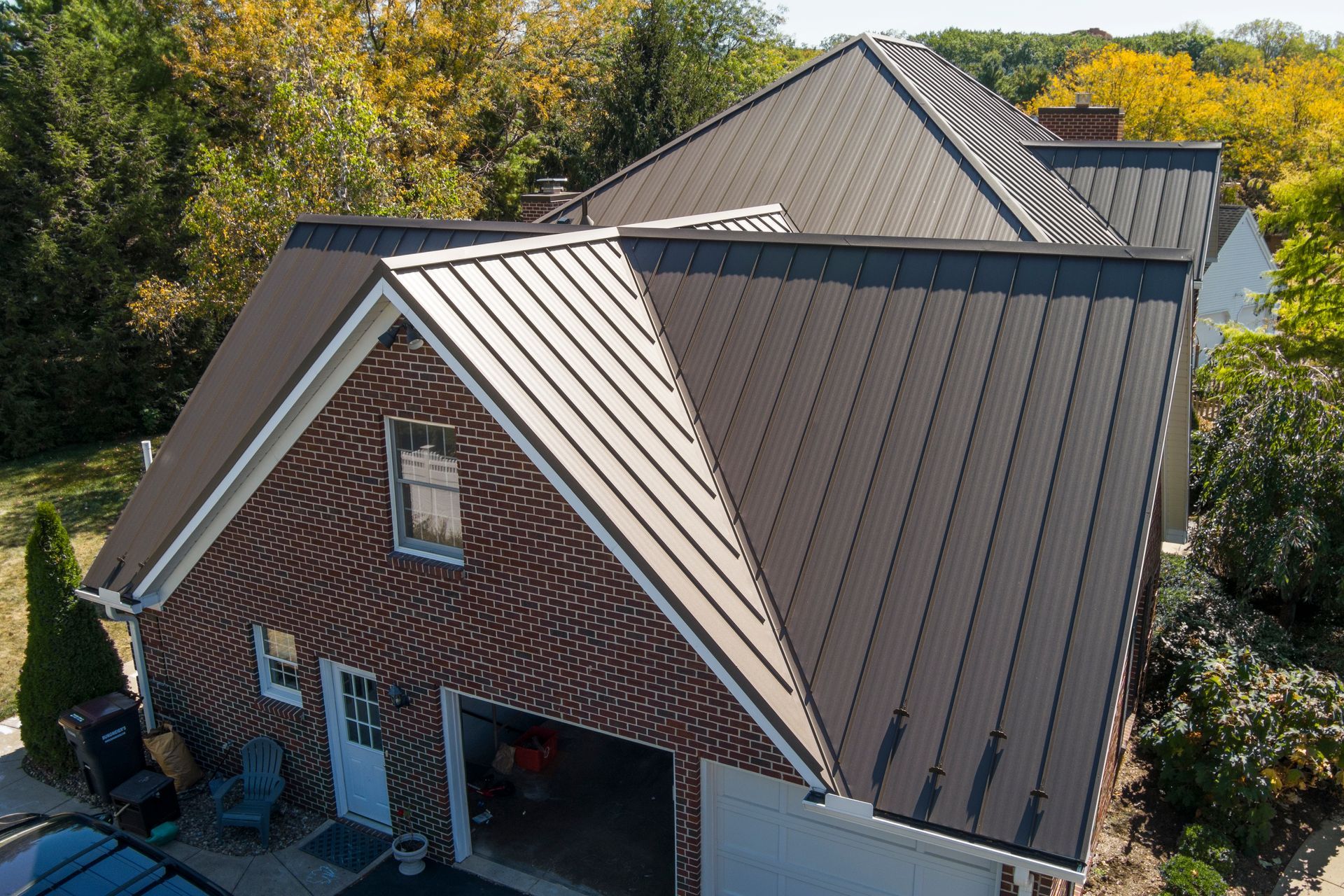 High-angle view of a brown metal roof on a brick home with an attached garage, surrounded by trees on a sunny day.