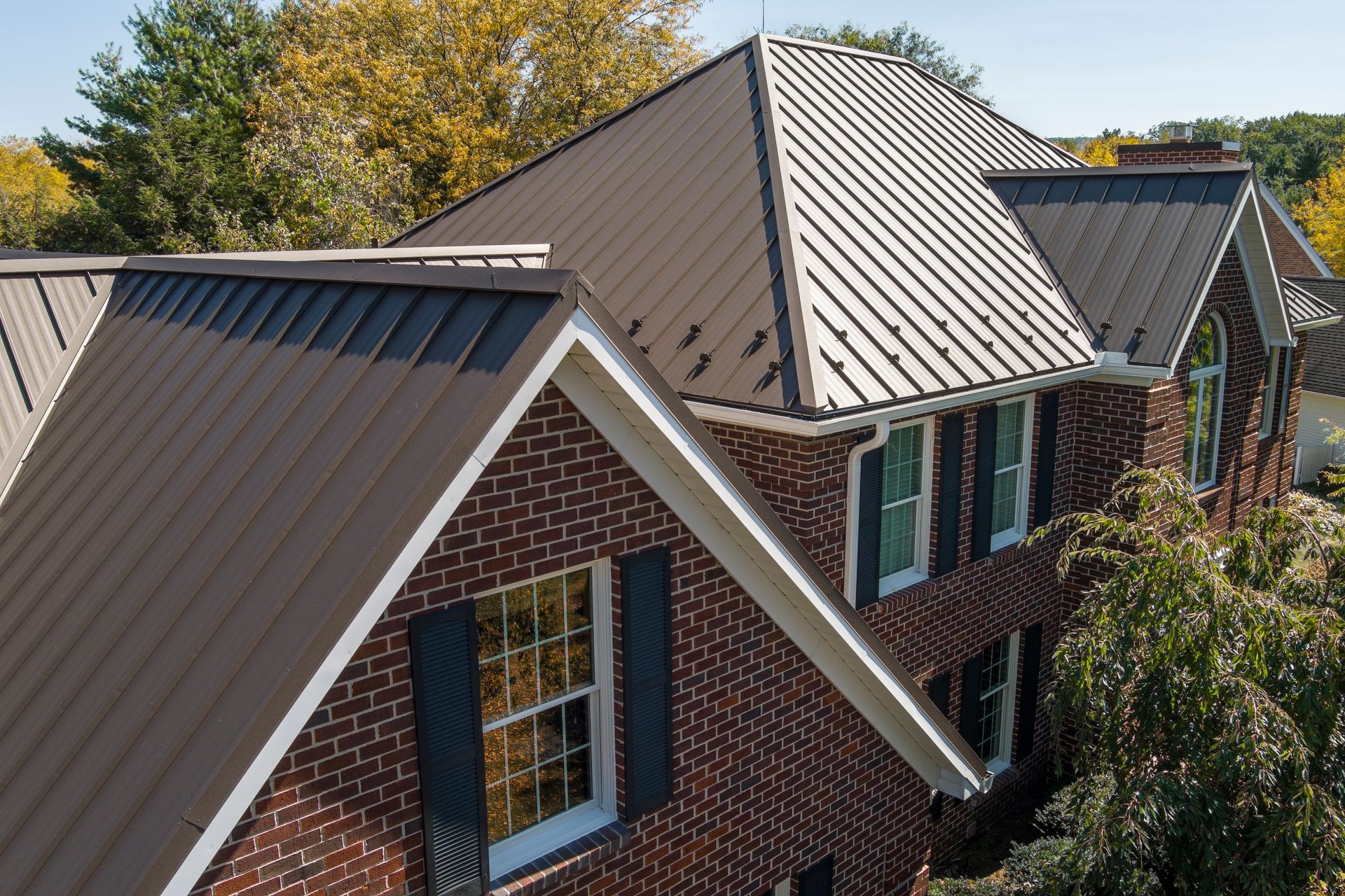 An elevated view of a brick house with a brown, standing-seam metal roof under a bright, clear blue sky.