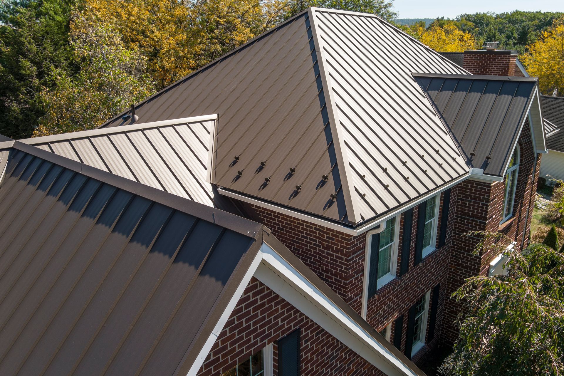 An aerial view of a brown metal standing-seam roof on a brick home, surrounded by trees on a sunny day.