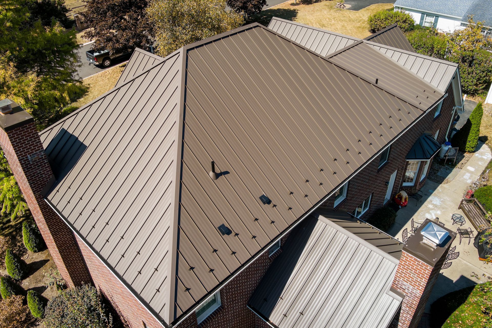 Aerial view of a residential brick home with a dark brown, patterned metal roof, surrounded by trees and a paved yard.