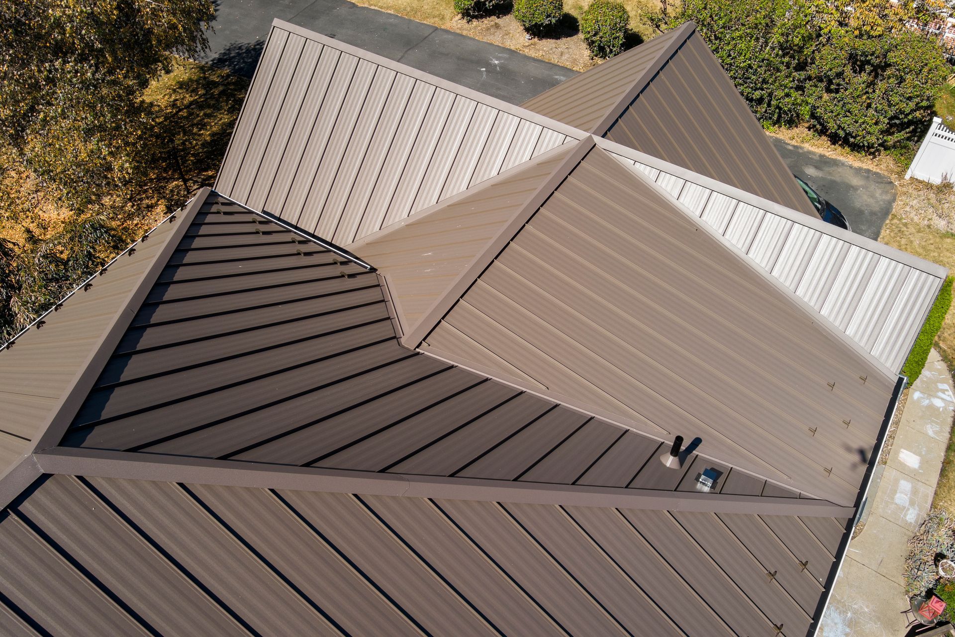 An overhead view of a dark brown metal roof with multiple intersecting planes and ridges on a sunny day.