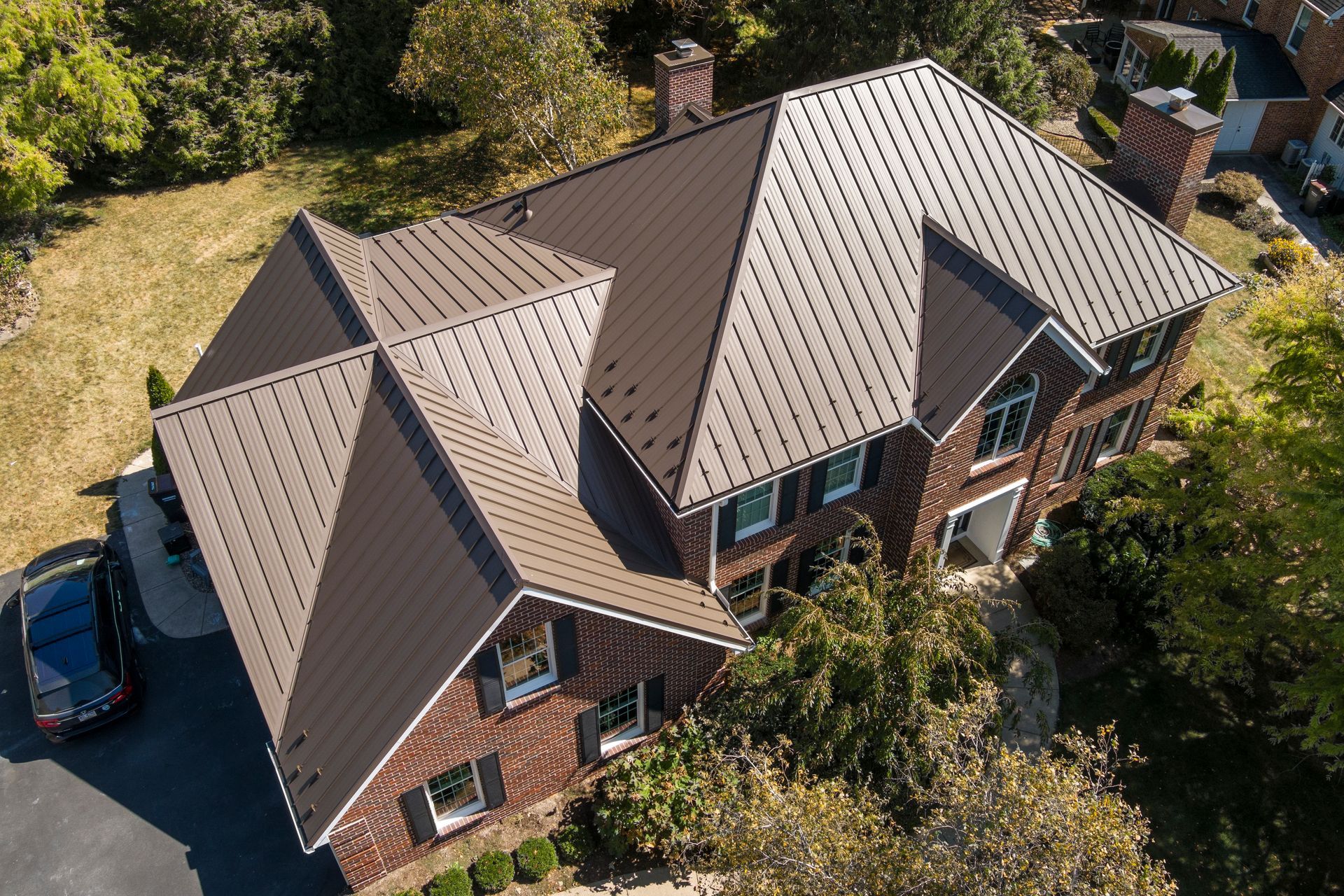 An aerial view of a brick house featuring a dark brown, multi-textured metal roof surrounded by trees and a driveway.