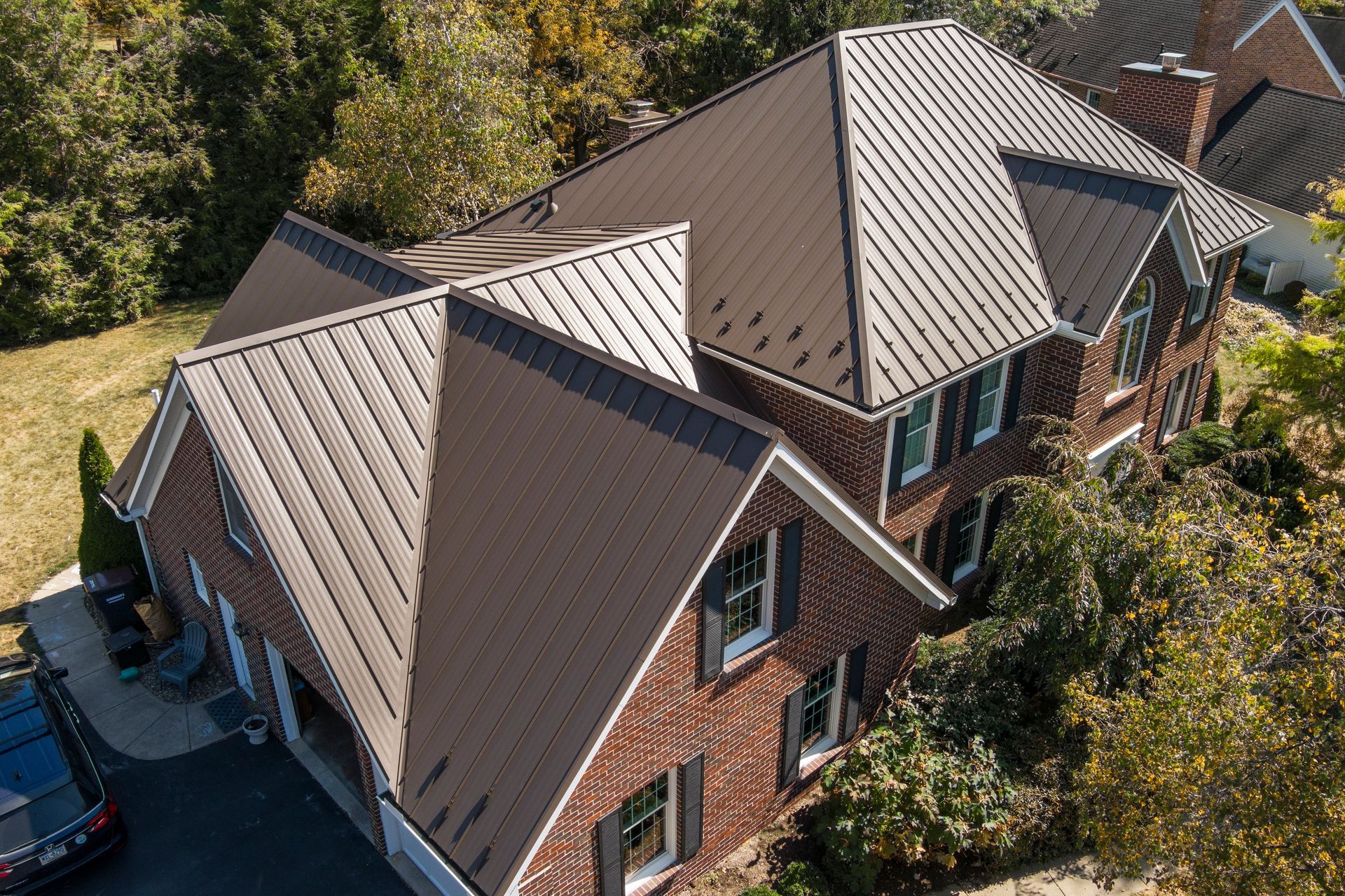 An aerial view of a brick house with a newly installed, dark brown metal roof, set in a sunny residential neighborhood.