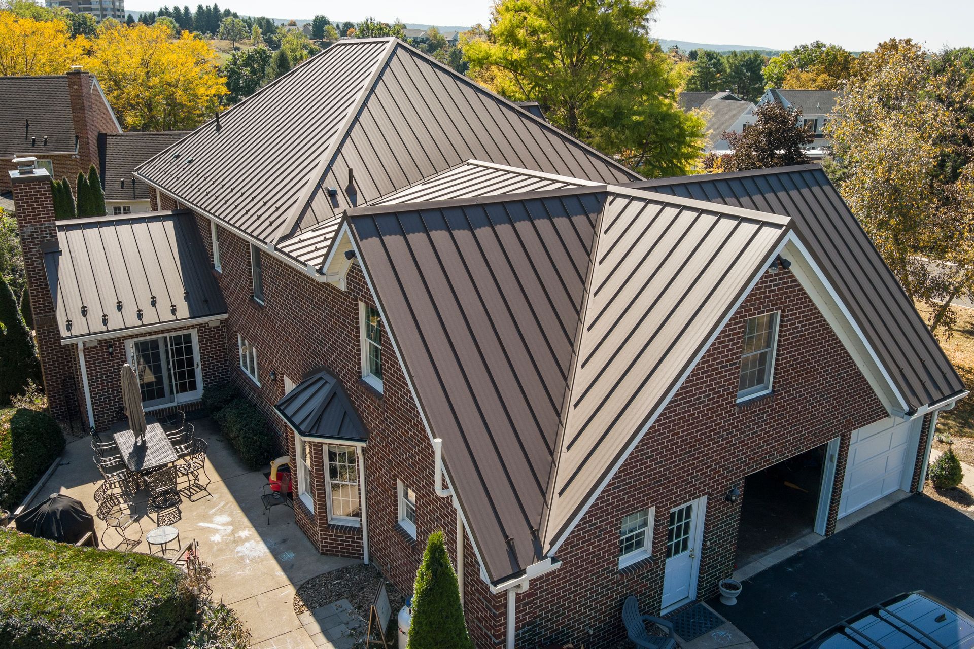 An aerial view of a brick house featuring a dark brown metal roof, with an attached garage and a backyard patio area.