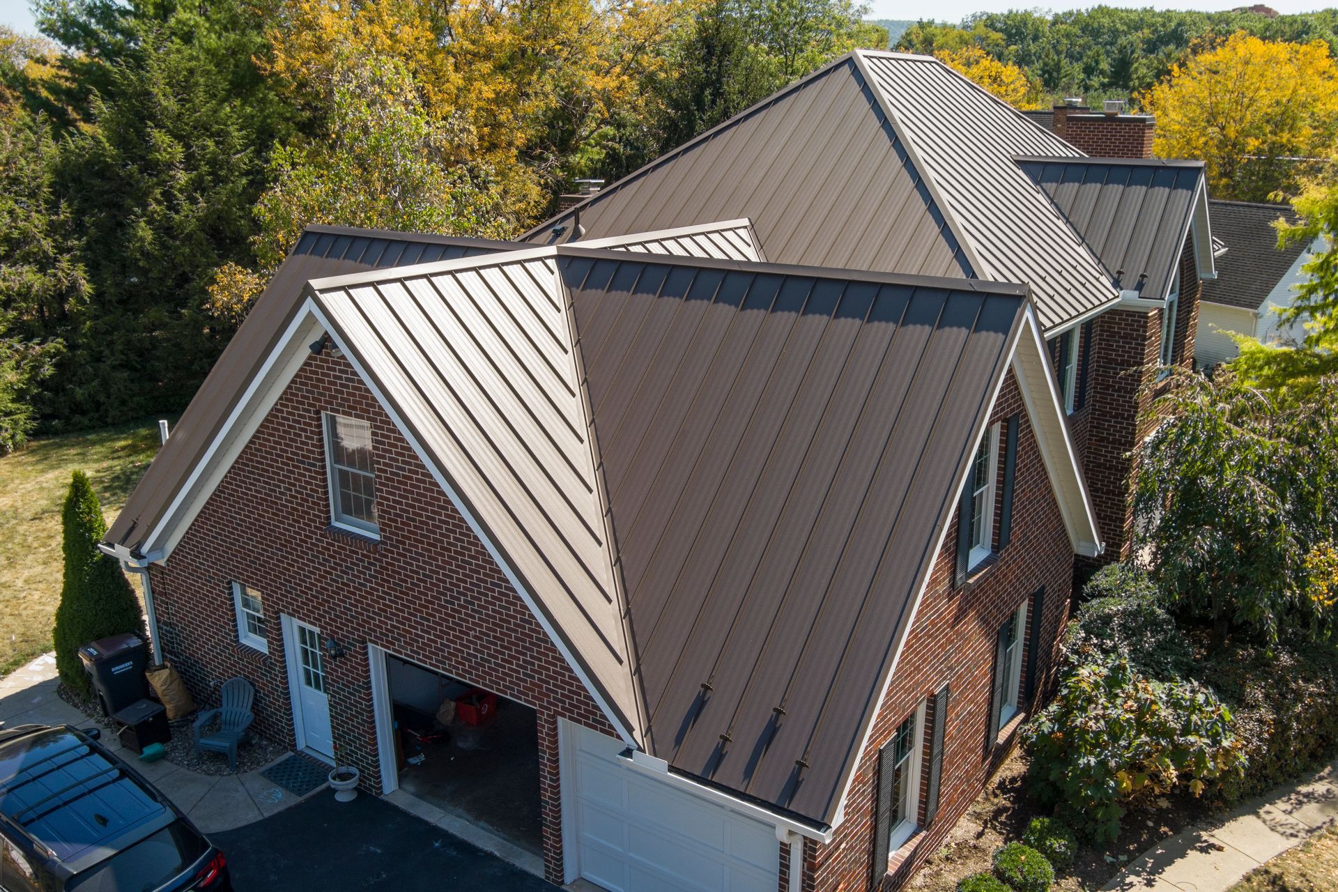 High-angle view of a brick house with a newly installed brown metal roof in a suburban setting surrounded by autumn trees.