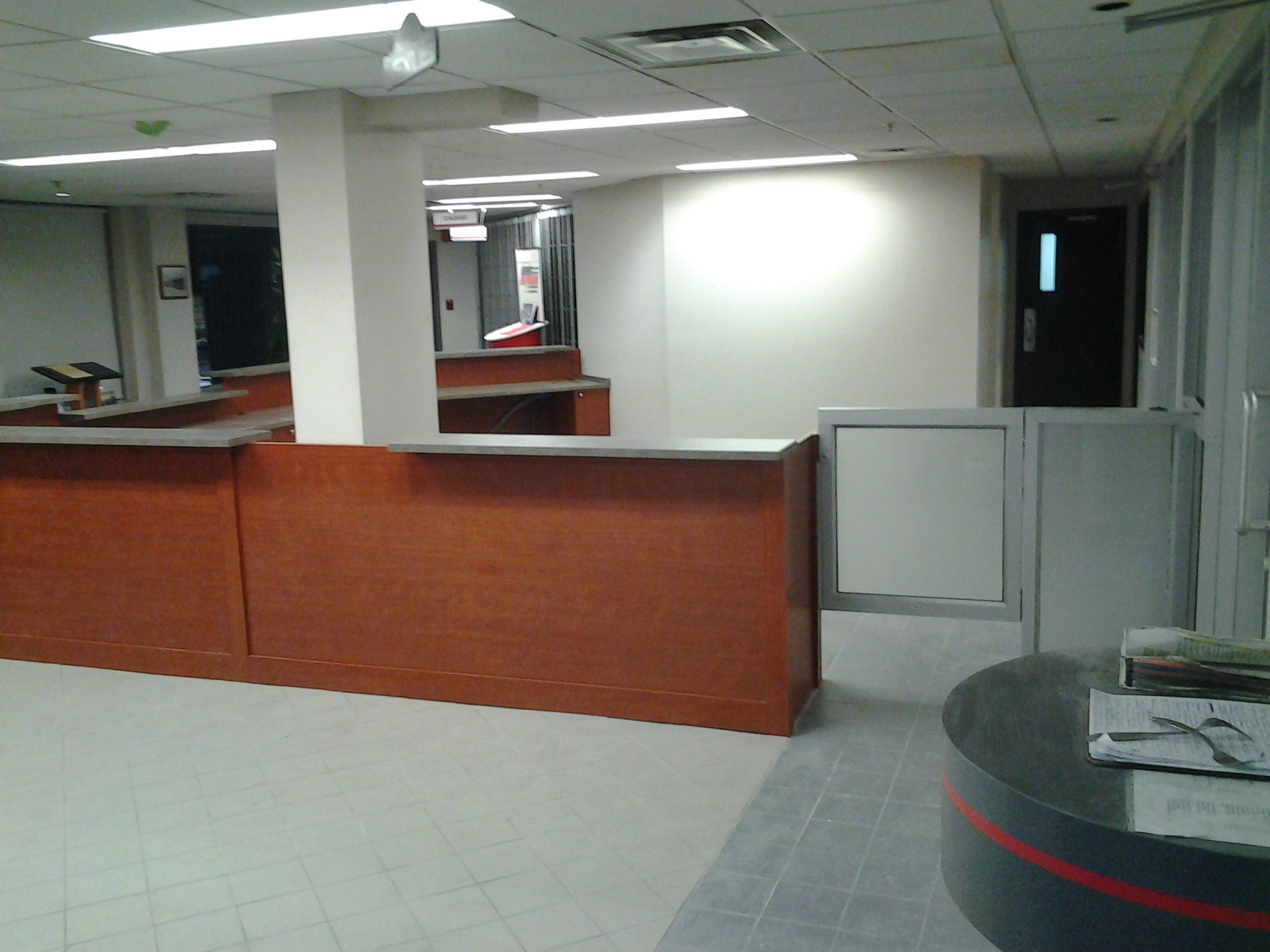 Intérieur du bureau : comptoirs d'accueil en bois, murs blancs, sol gris, porte noire, éclairage vif.