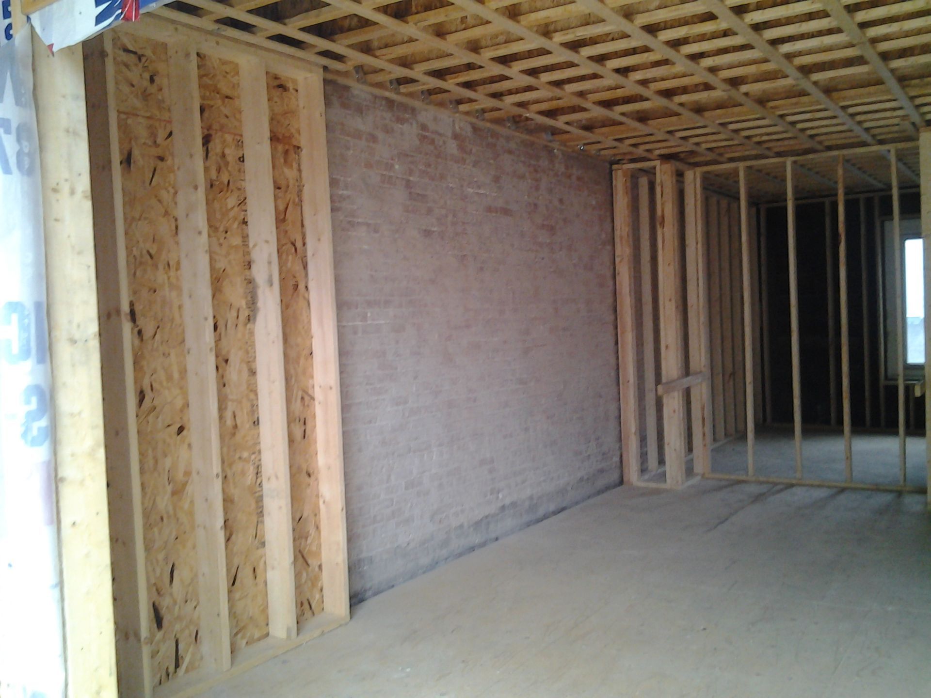 Intérieur d'un bâtiment en construction, montrant une charpente en bois, un sol en béton et un mur en briques.