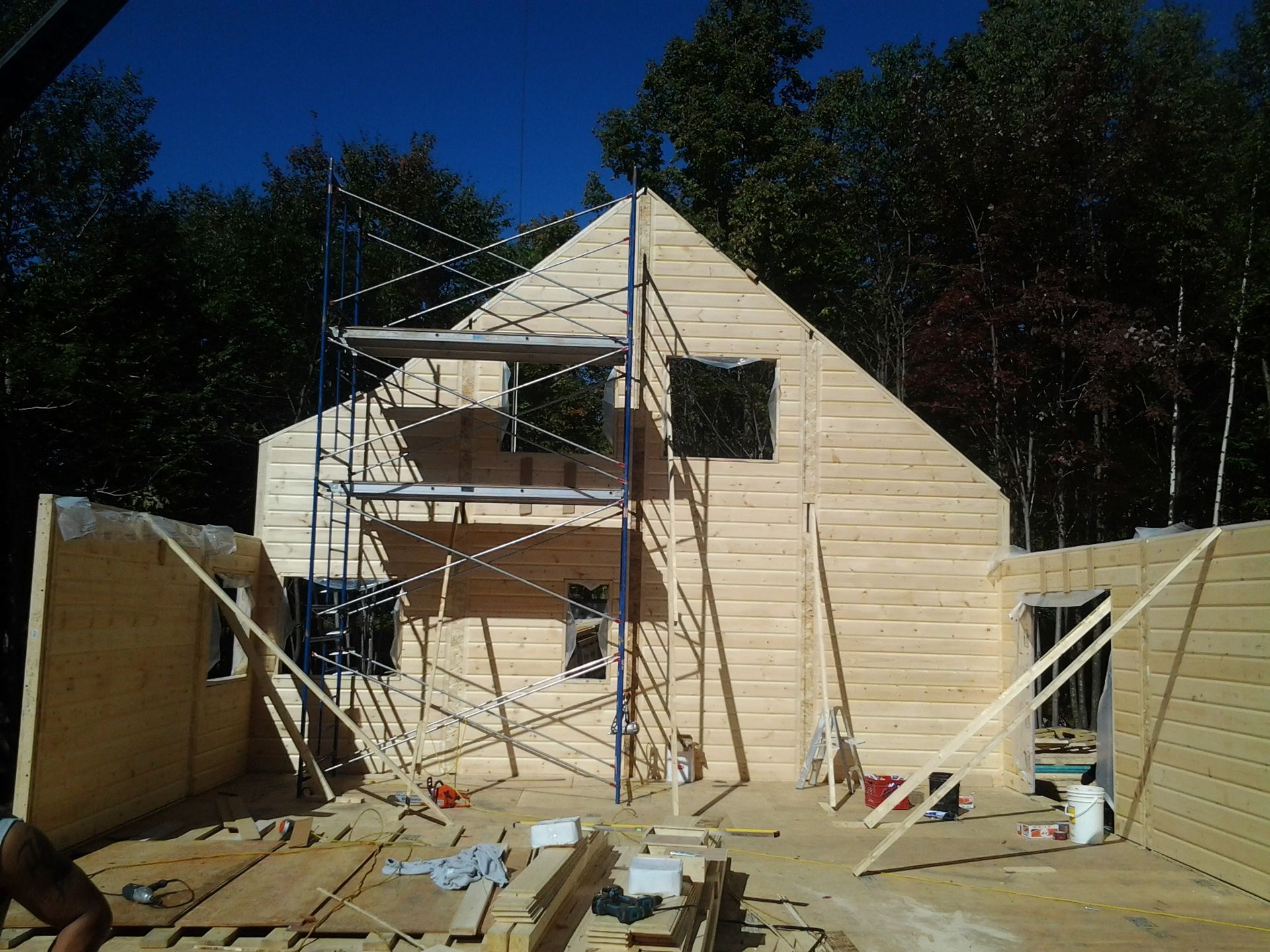 Construction d'une maison en bois avec échafaudages, sous un ciel bleu, entourée d'arbres.