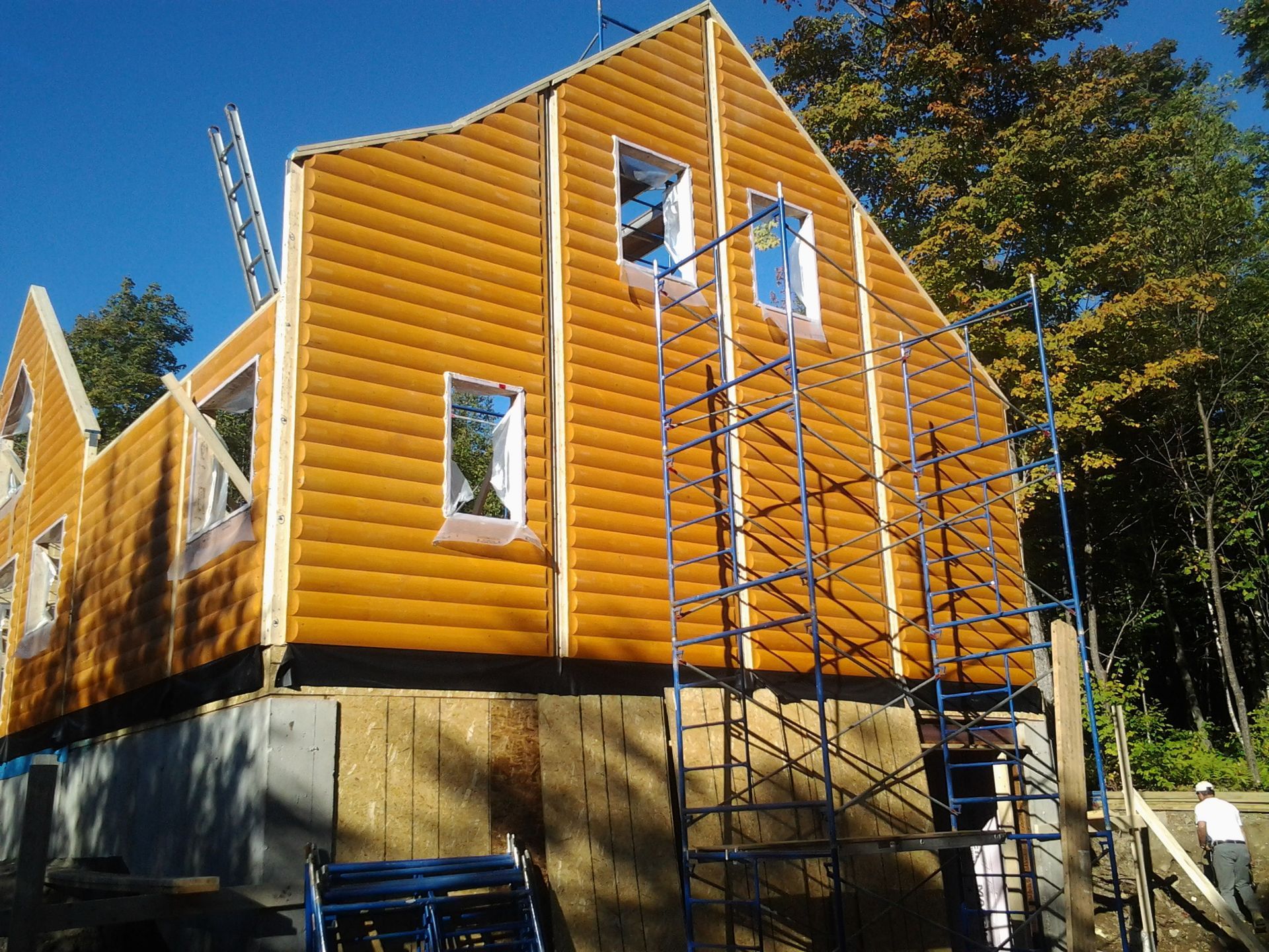 Maison en construction, vue extérieure ; bardage marron, échafaudages, fenêtres et arbres.