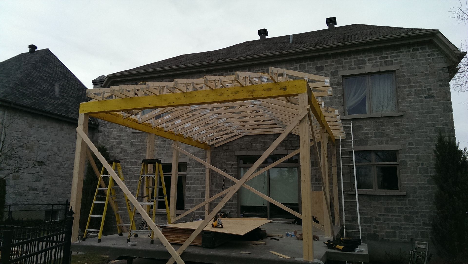 Structure de pergola en bois en construction, adossée à une maison en pierre.