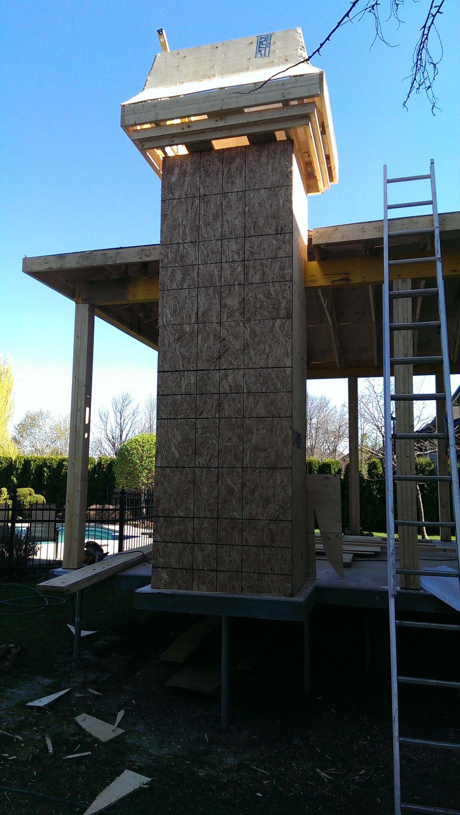 Construction d'une structure haute, revêtue de panneaux de bois et dotée d'une extension de toit, à côté d'un bâtiment et d'une échelle, à l'extérieur.