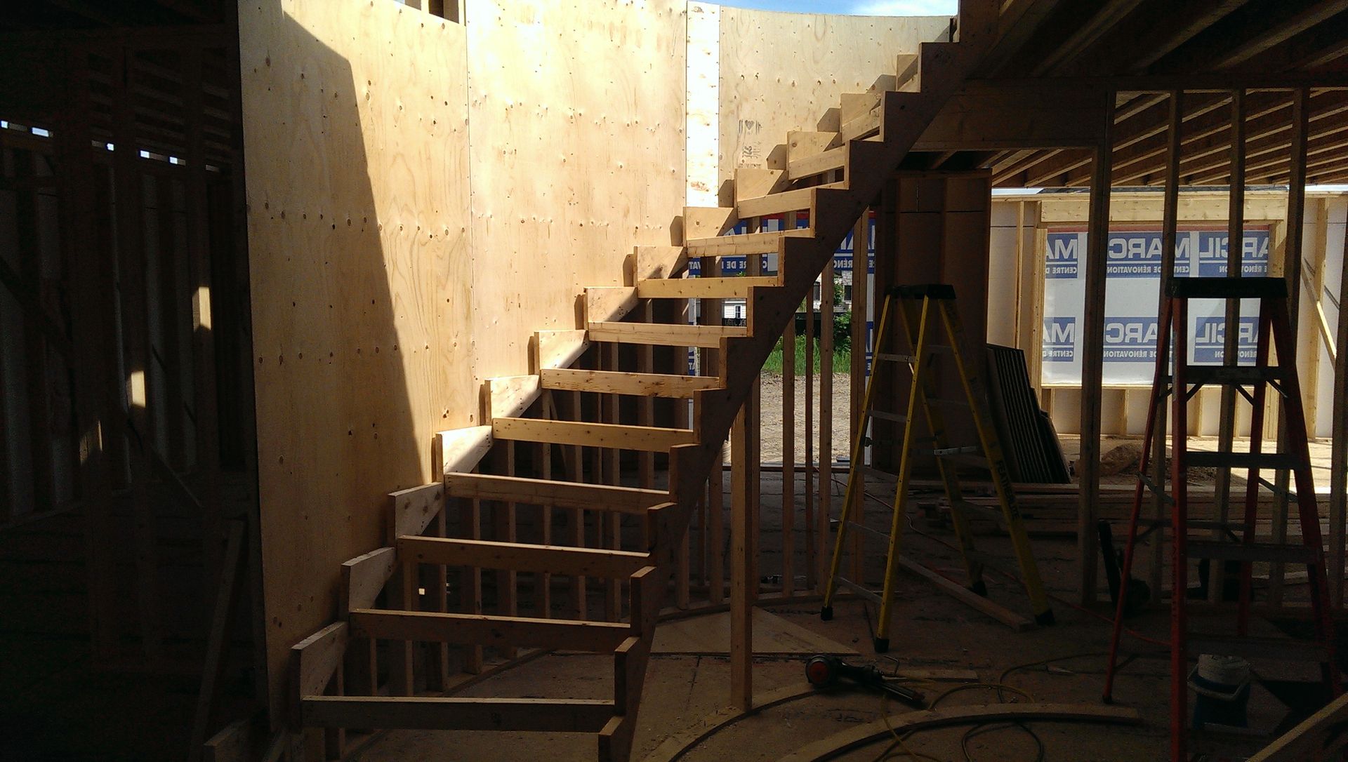 Escalier en bois en construction à l'intérieur d'un bâtiment ; la lumière pénètre par une fenêtre voisine.
