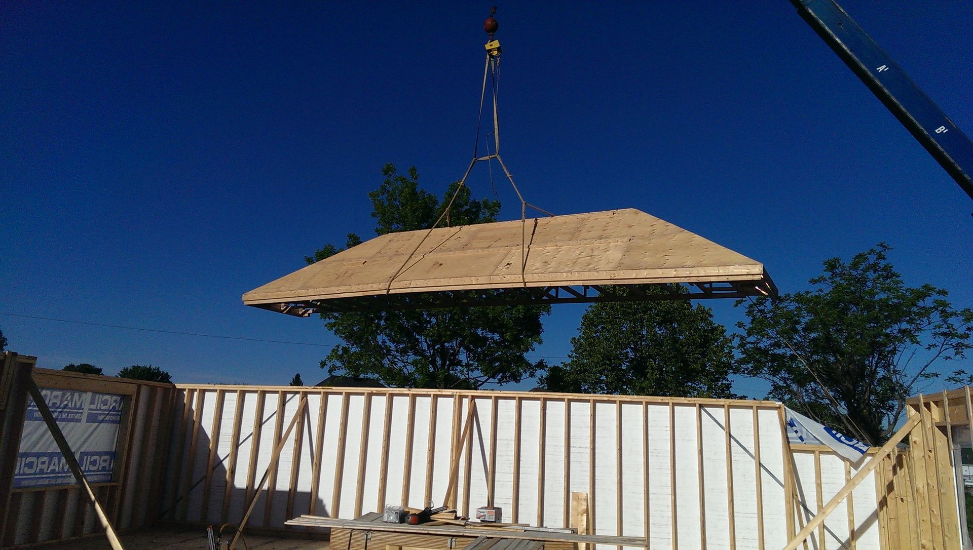 Une section de toiture soulevée par une grue au-dessus d'un bâtiment en construction, sur fond de ciel bleu.