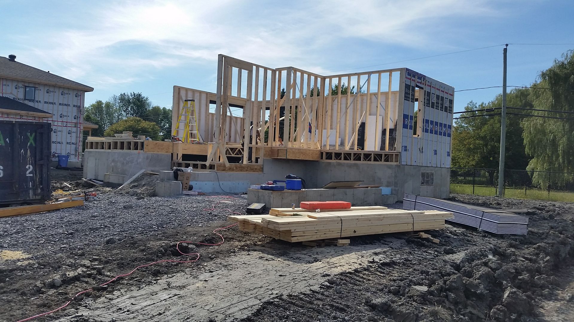 Une maison en construction ; une ossature en bois sur des fondations en béton, entourée de matériaux de construction, et le ciel est bleu.