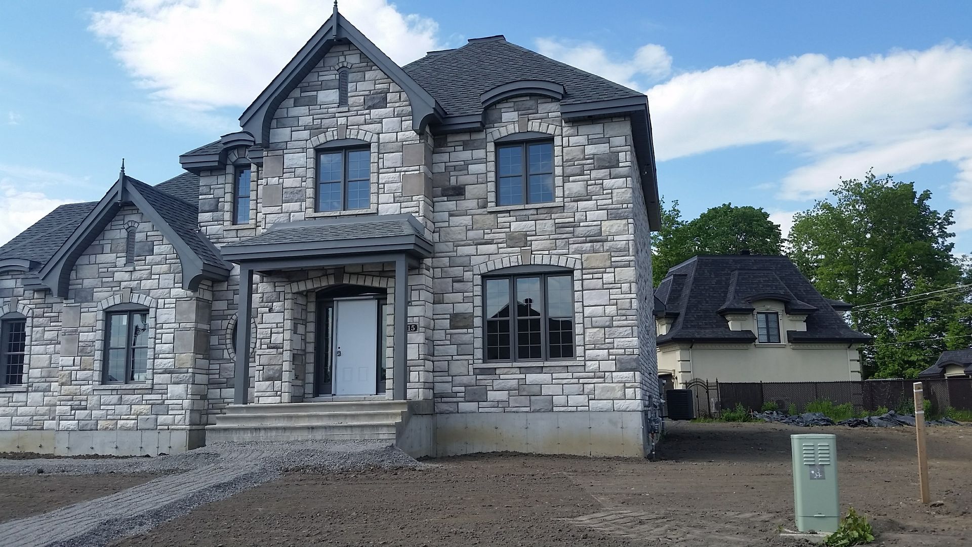 Maison à deux étages revêtue de pierre, avec des fenêtres à encadrement noir et un bâtiment indépendant en arrière-plan, sur fond de ciel bleu.