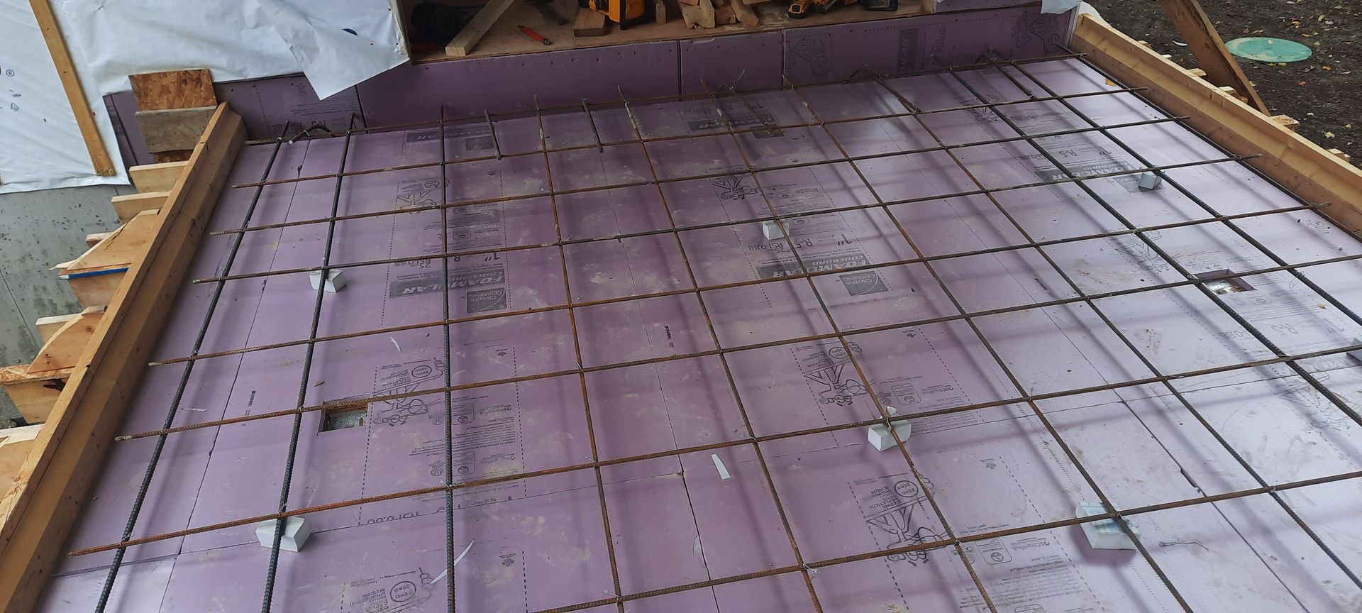 Grille d'armature métallique posée sur un panneau isolant violet à l'intérieur d'un cadre en bois, probablement pour un coulage de béton.