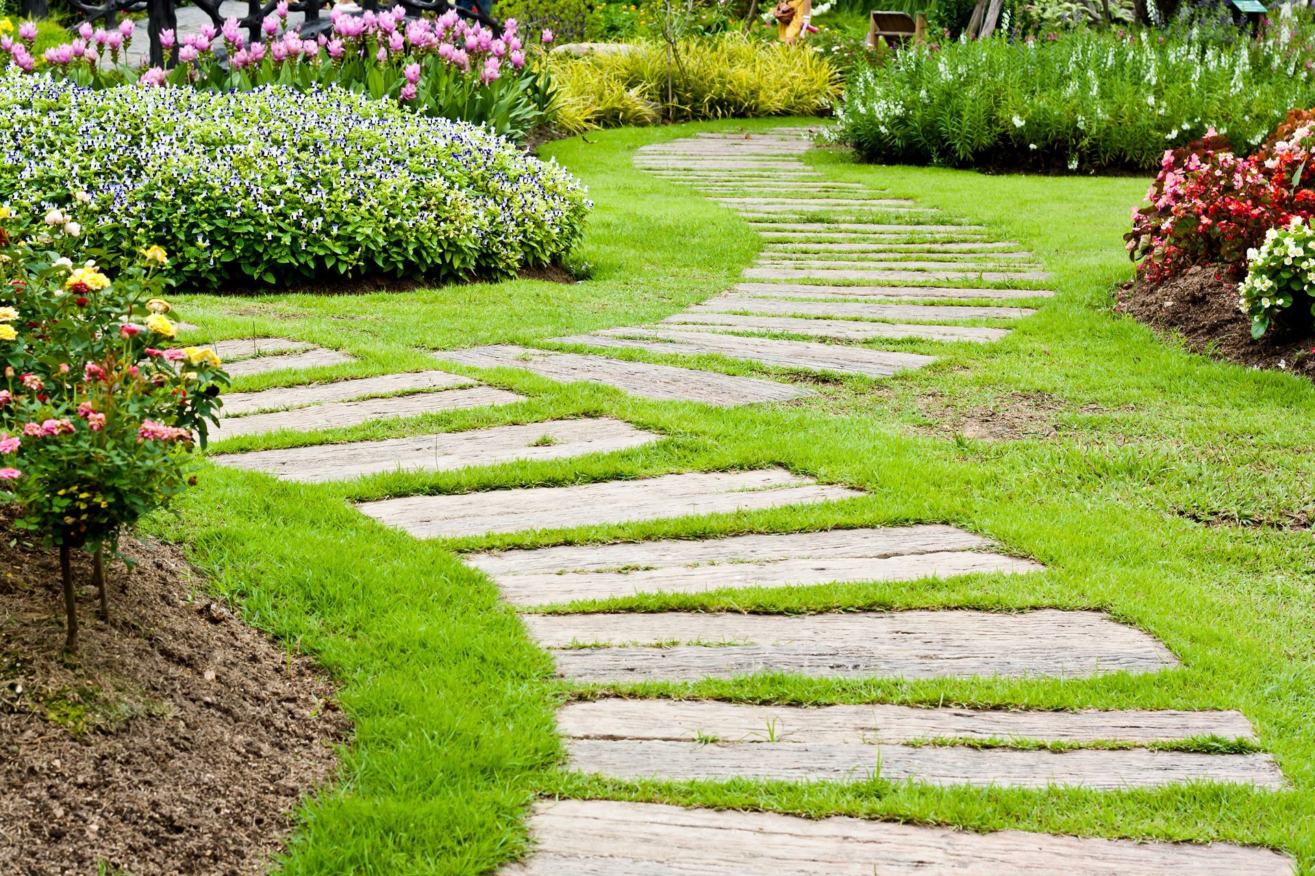 A stone walkway in a garden surrounded by grass and flowers