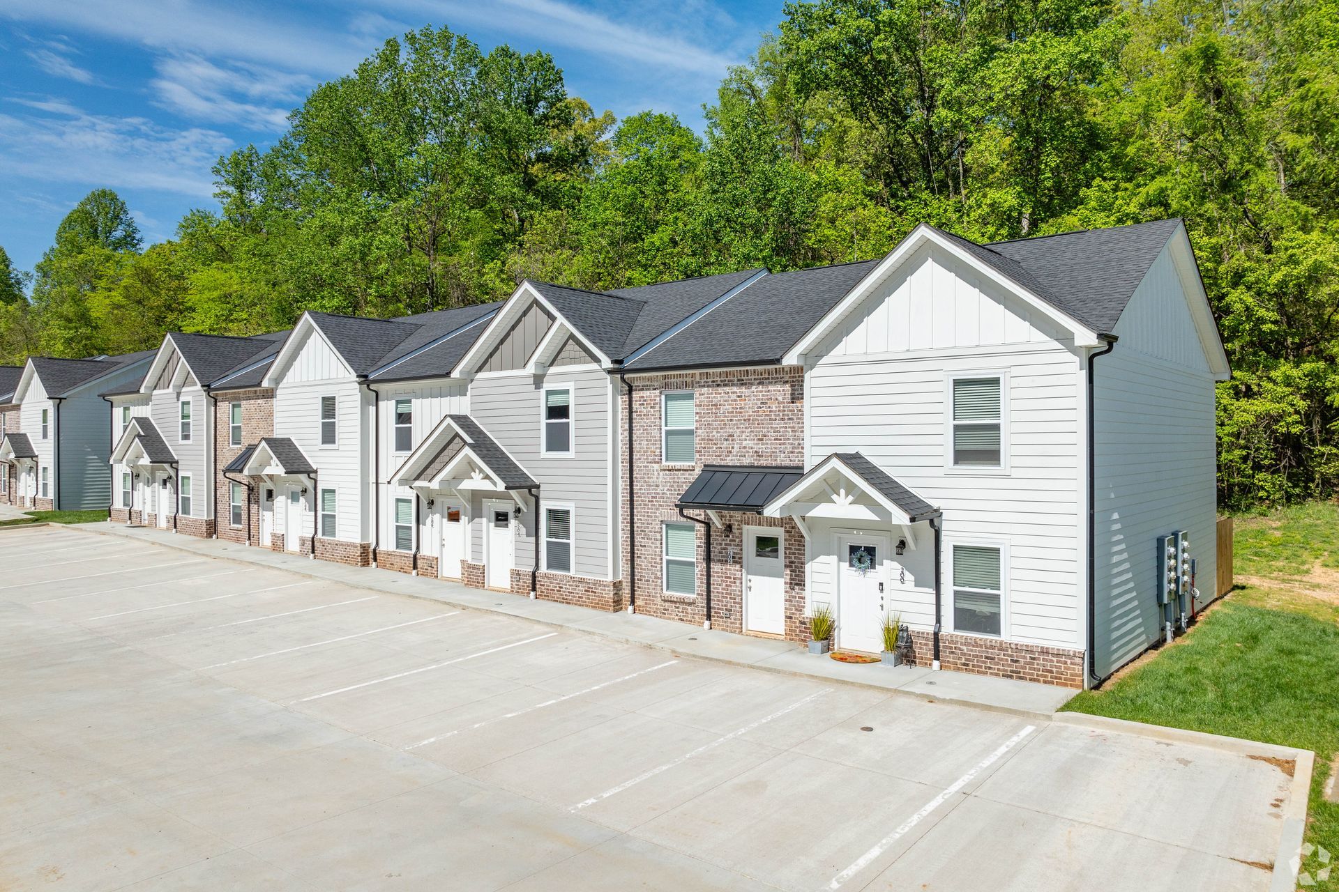 Row of townhouses with gray and white exteriors, asphalt parking, green trees in background.
