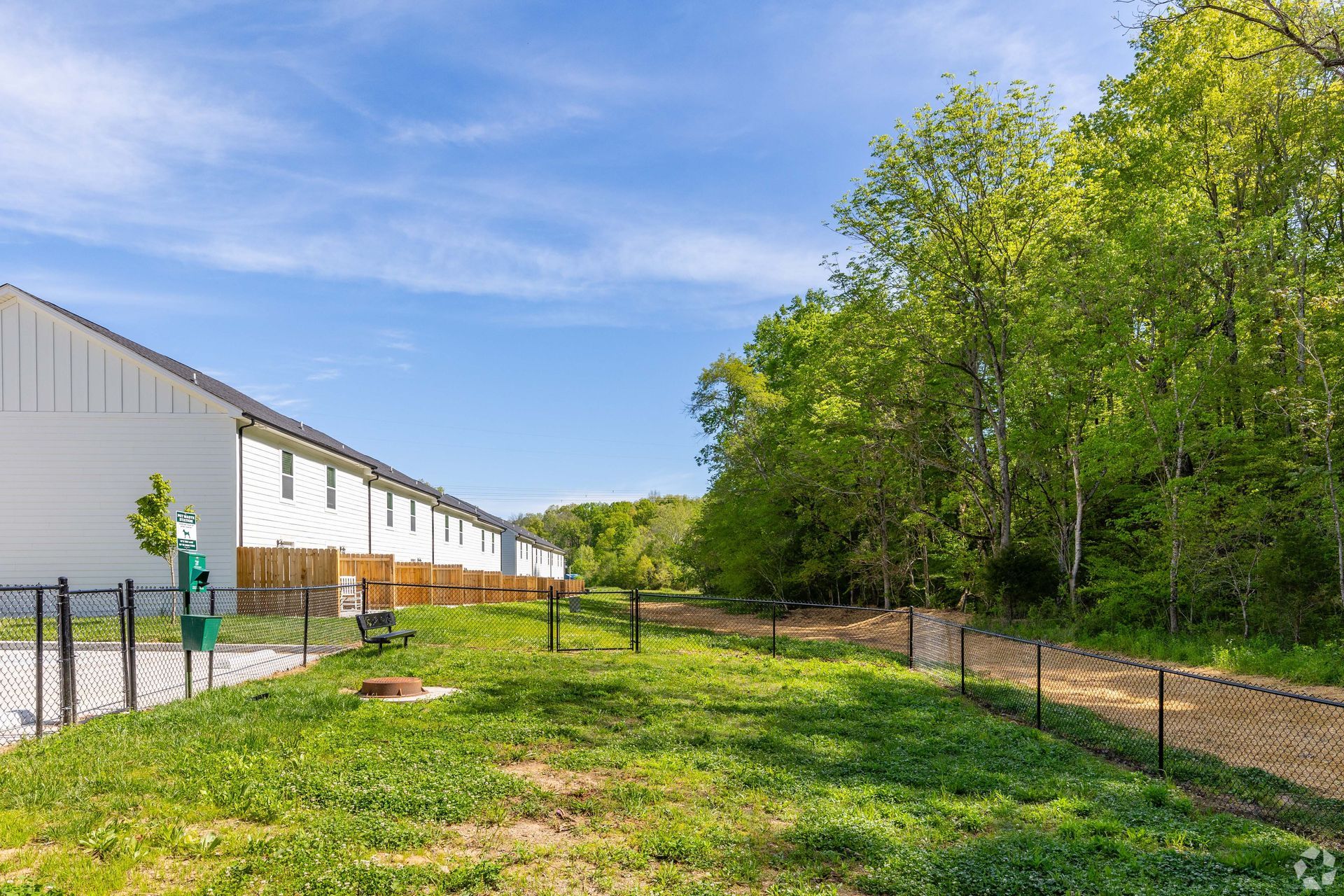 Dog park with green grass, trees, and row of white townhomes in background.