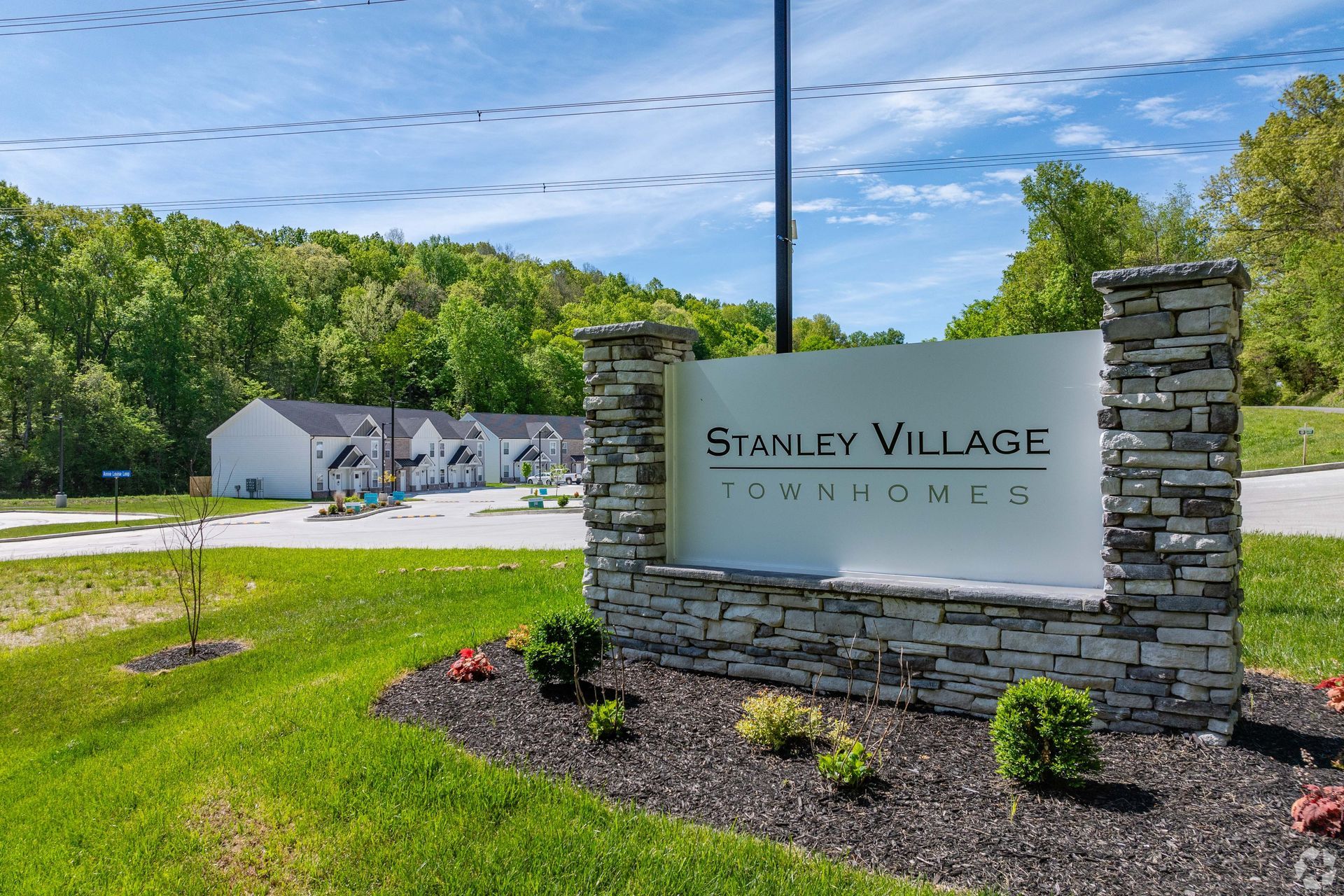 Sign for Stanley Village Townhomes with townhouses in the background, set against a green, wooded area.