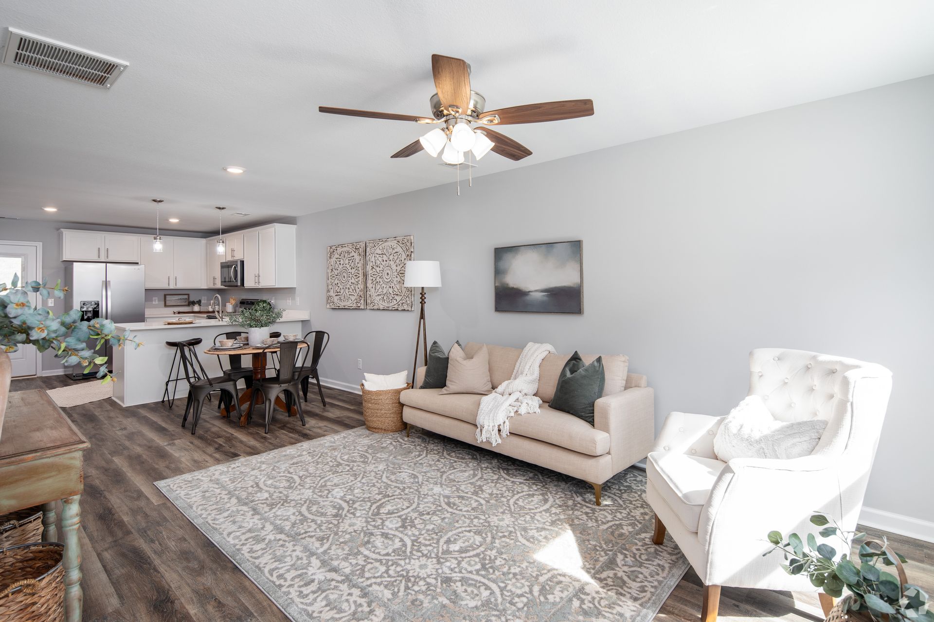 Living room with light gray walls, open to a kitchen. Beige sofa, white armchair, area rug, and wooden ceiling fan.