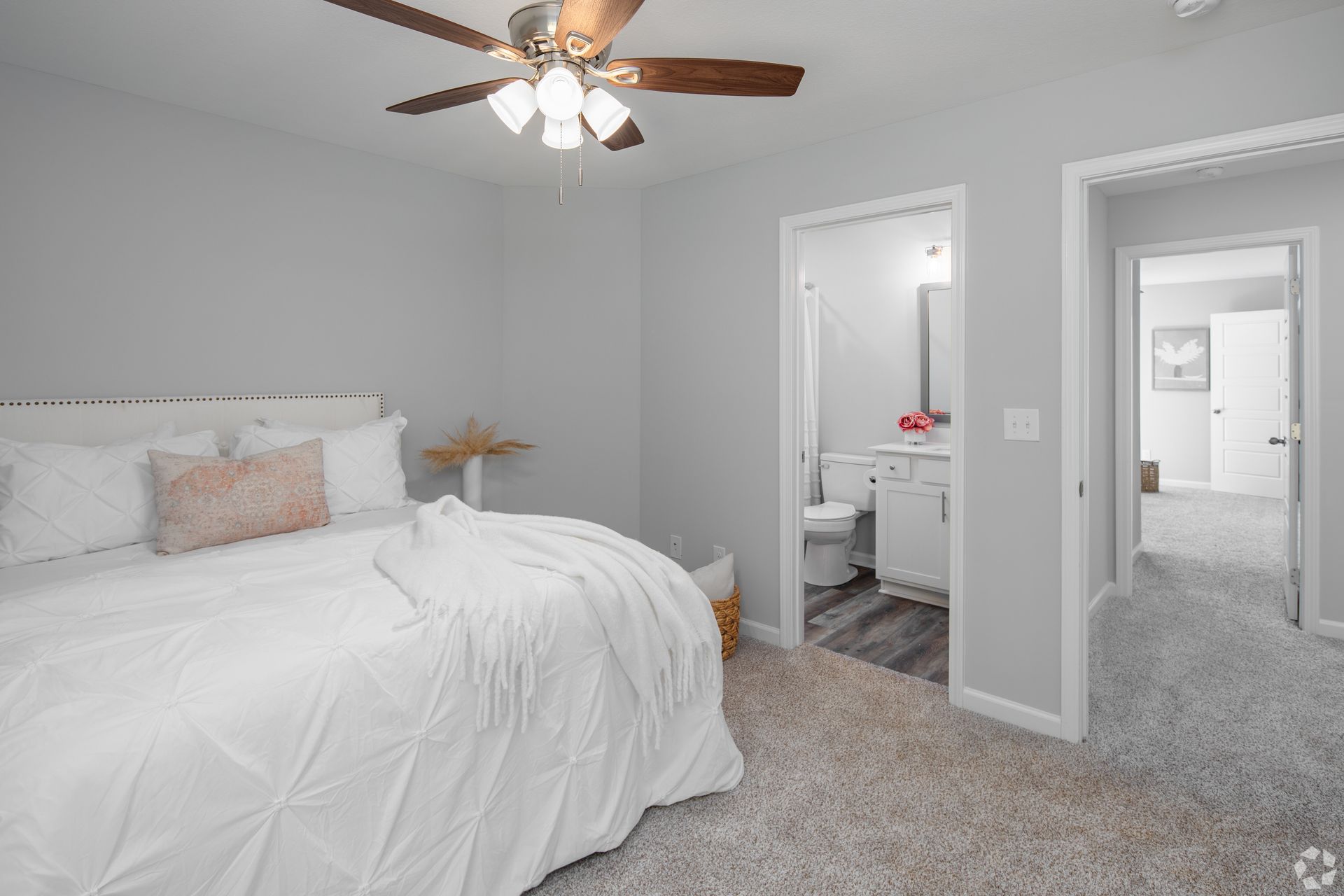 Bedroom with bed, bathroom, and doorway. Light gray walls, white bedding, ceiling fan, and carpet.