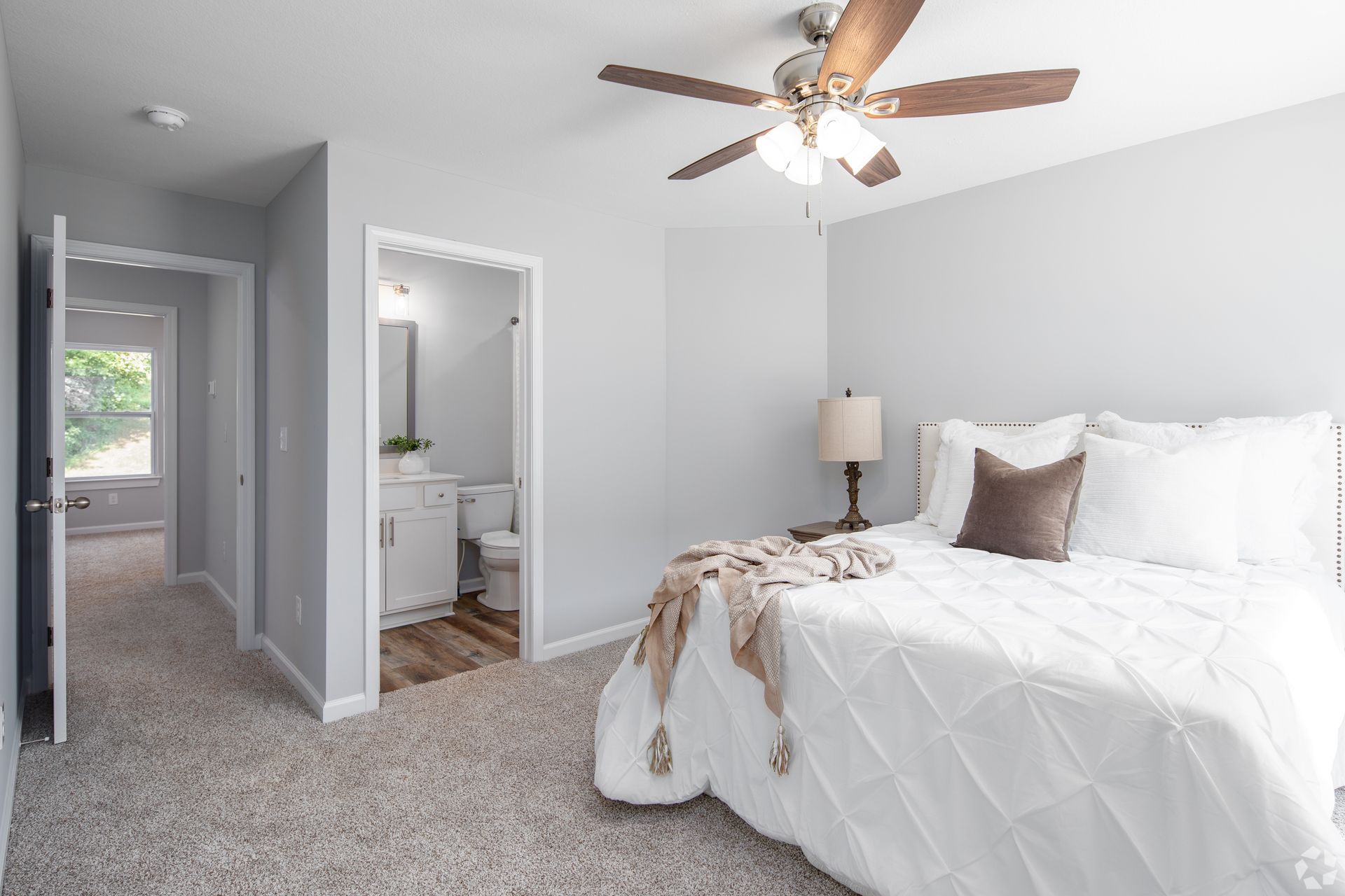 Bedroom with a white bed, beige carpet, and light gray walls. A bathroom is visible in the doorway.