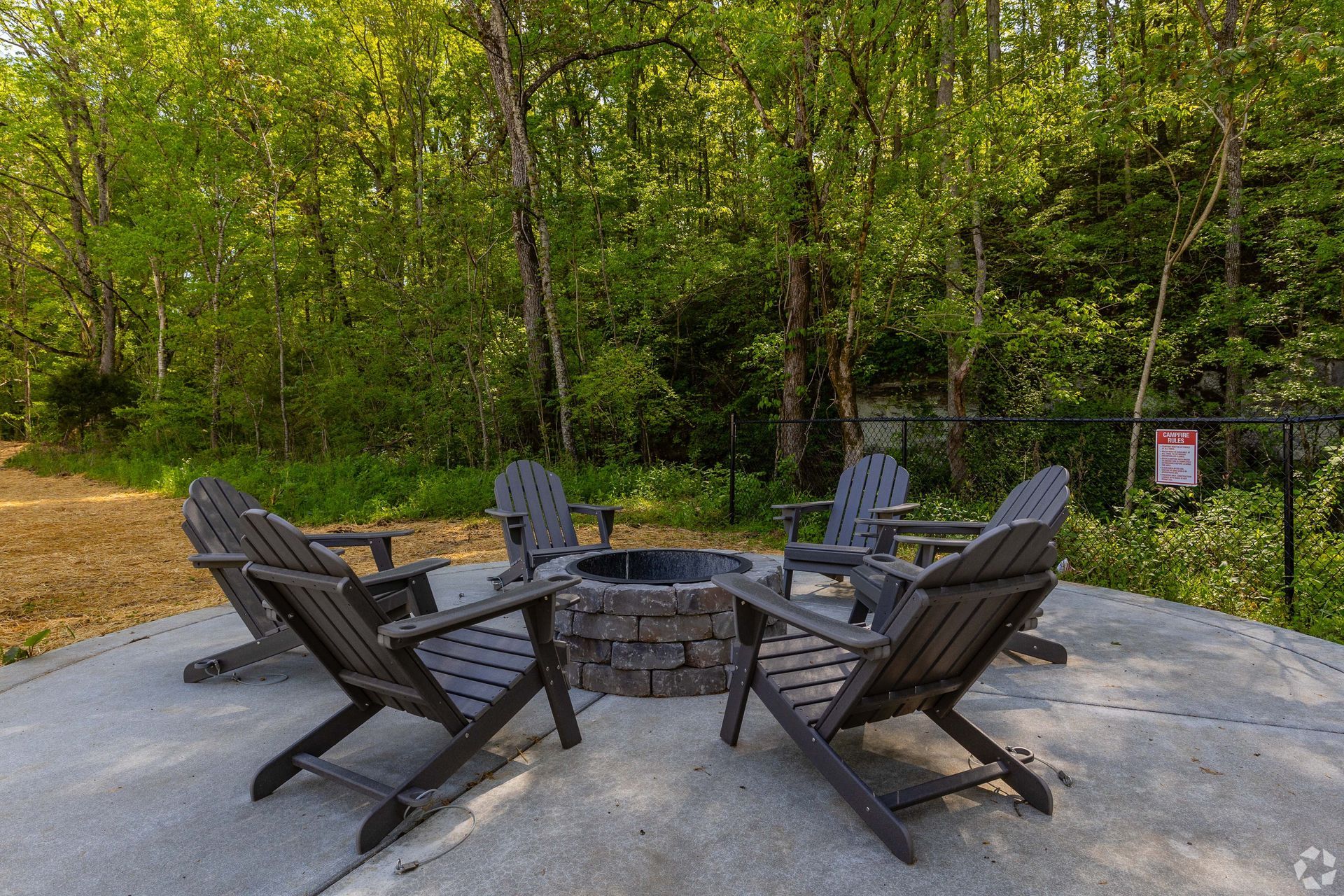 Four wooden chairs arranged around a stone fire pit on a concrete patio, surrounded by trees.