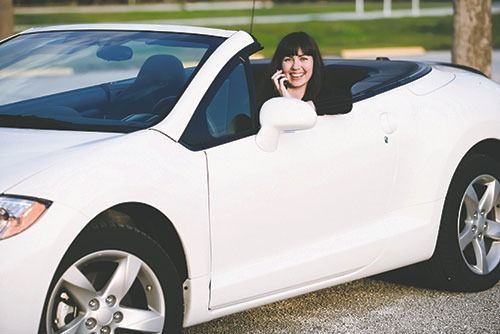 Woman in a white convertible smiles while talking on a cell phone.