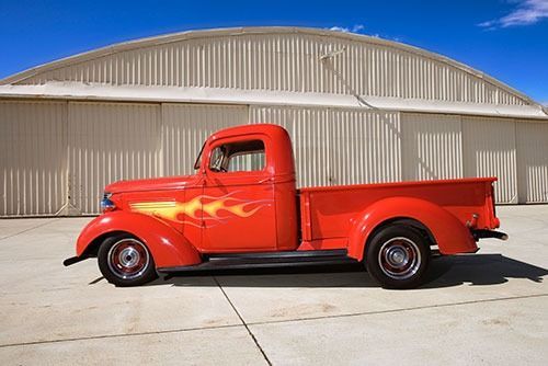 Red vintage pickup truck with flame decals, parked in front of a tan hangar on a clear day.