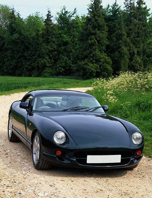 Dark green sports car parked on a dirt road, trees in the background.