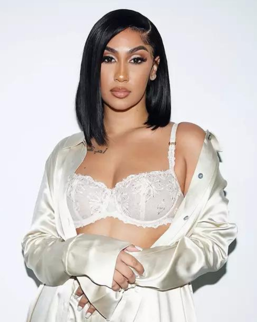 A woman in a bra and jacket is standing in front of a white wall.